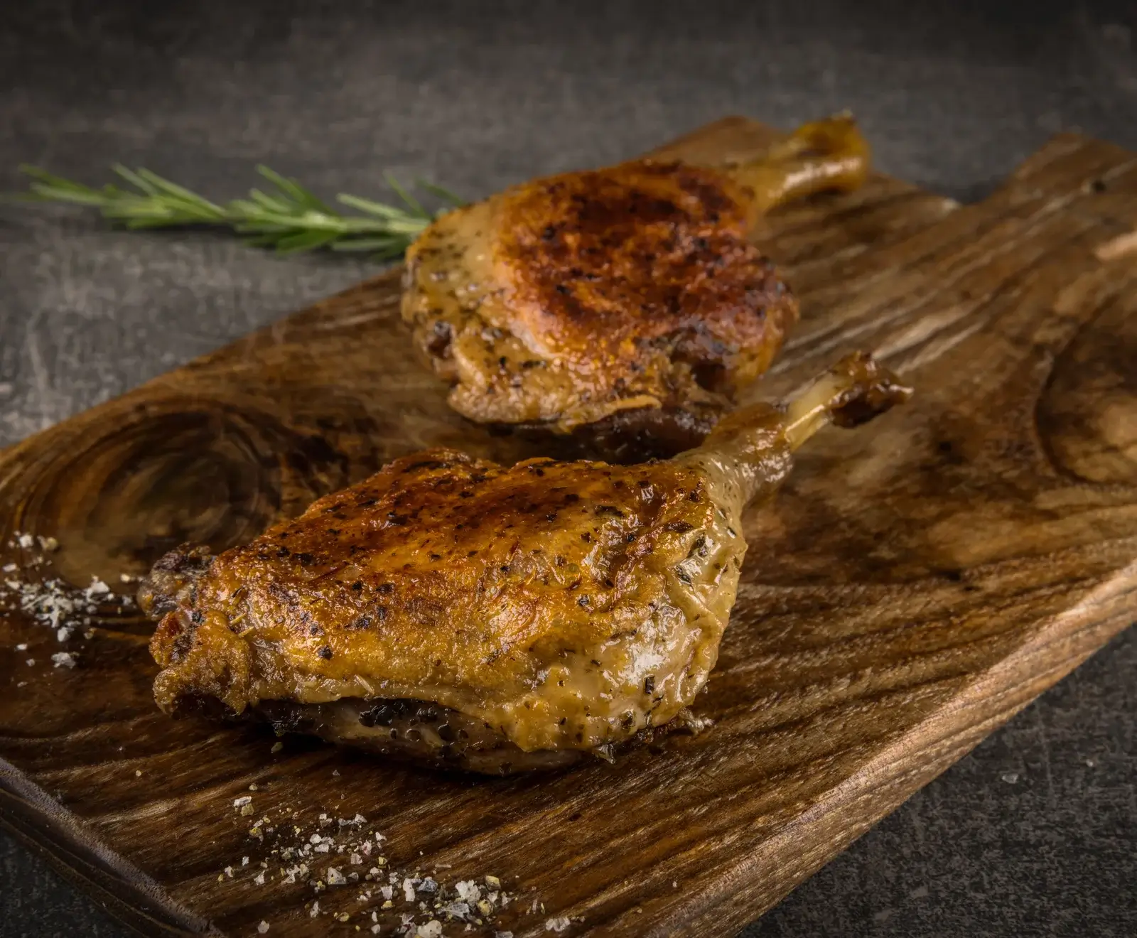 Zwei knusprig gebratene Entenkeulen mit goldbrauner Haut, gewürzt mit Kräutern und Pfeffer, liegen auf einem dunklen Holzbrett. Grobes Salz und Pfeffer sind verstreut, ein Rosmarinzweig liegt im Hintergrund.