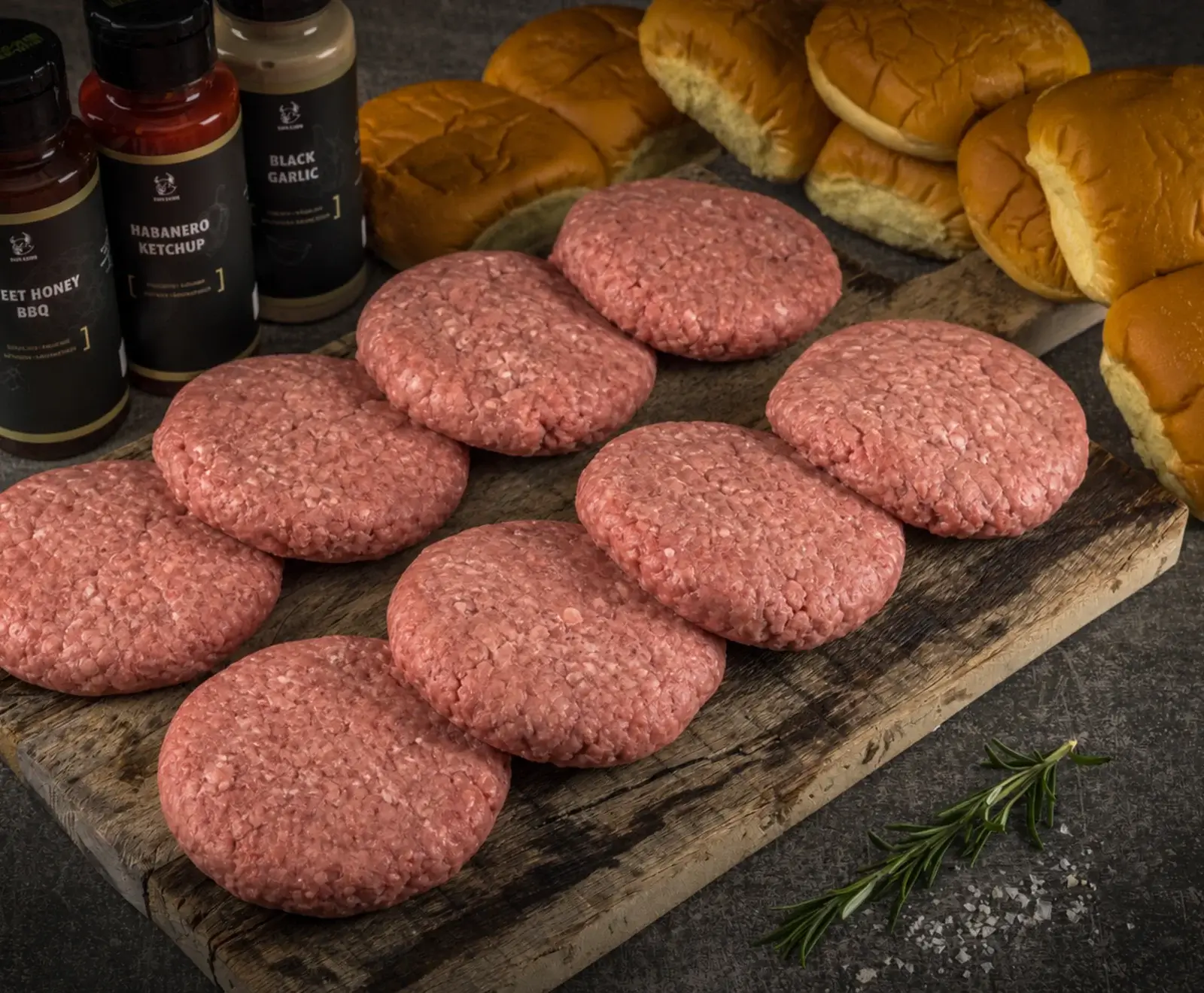 Rohe Burger-Patties auf einem Holzbrett, umgeben von Burger-Brötchen, Saucenflaschen (Sweet Honey BBQ, Habanero Ketchup, Black Garlic), Rosmarin und grobem Salz.