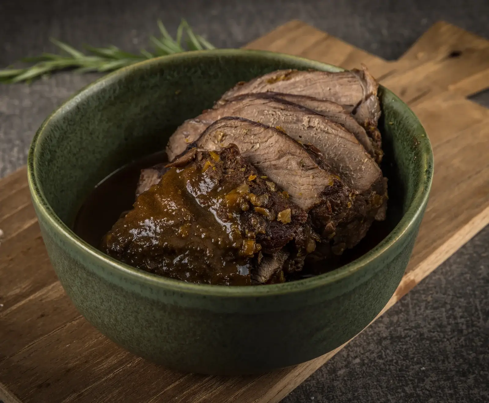 Nahaufnahme von saftigem, in Scheiben geschnittenem Braten mit reichhaltiger Bratensoße in einer dunkelgrünen Keramikschale, auf einem rustikalen Holzbrett. Ein Rosmarinzweig liegt im Hintergrund.