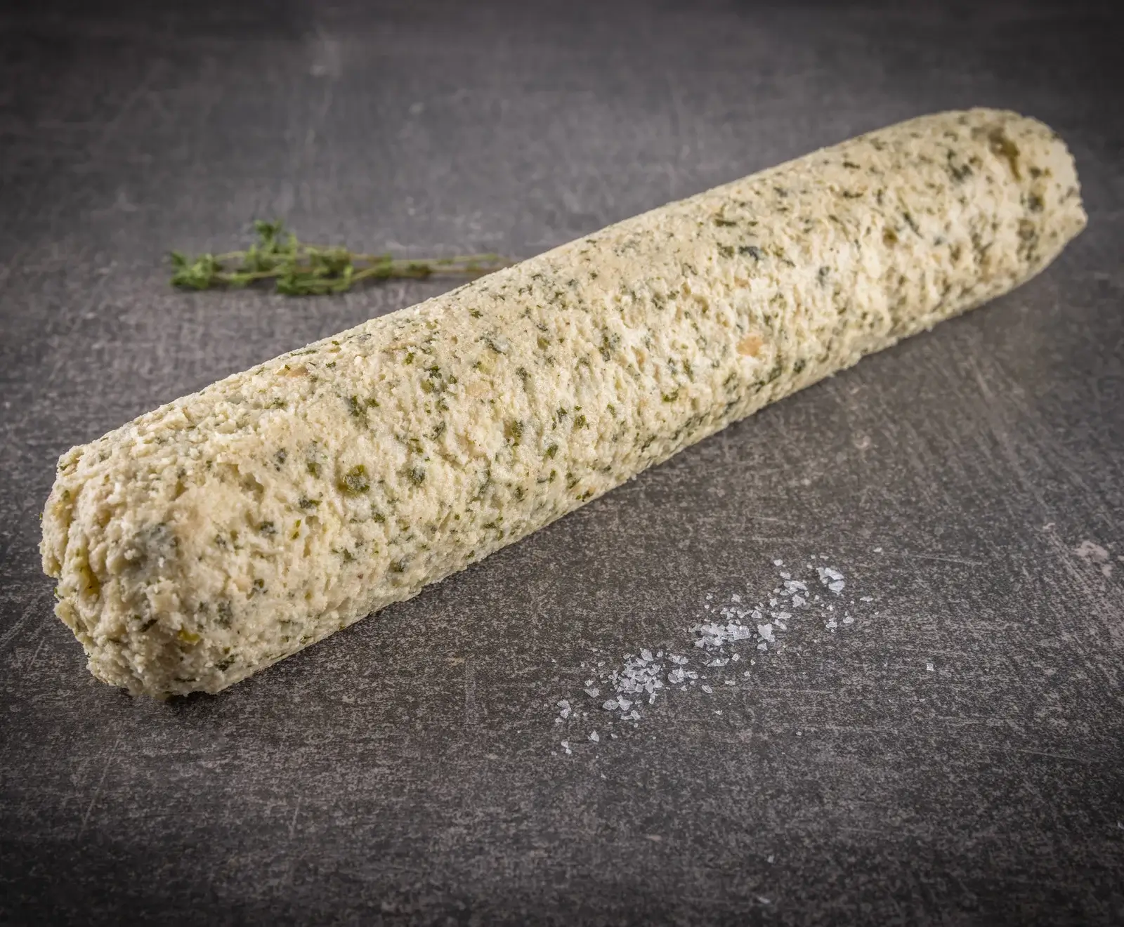 Ein zylindrisches Stück Kräuterbutter mit grünen Kräuterstücken liegt auf einer dunklen, strukturierten Oberfläche. Im Hintergrund sind frische Thymianzweige, im Vordergrund grobe Salzkristalle verstreut.