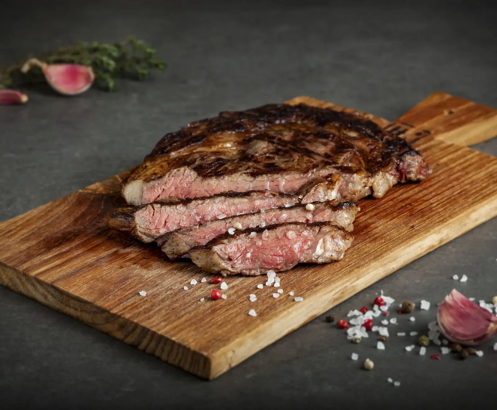 Ein perfekt gegrilltes, in Scheiben geschnittenes Steak mit rosa Kern auf einem rustikalen Holzbrett, garniert mit grobem Salz und bunten Pfefferkörnern. Knoblauch und Thymian im Hintergrund.