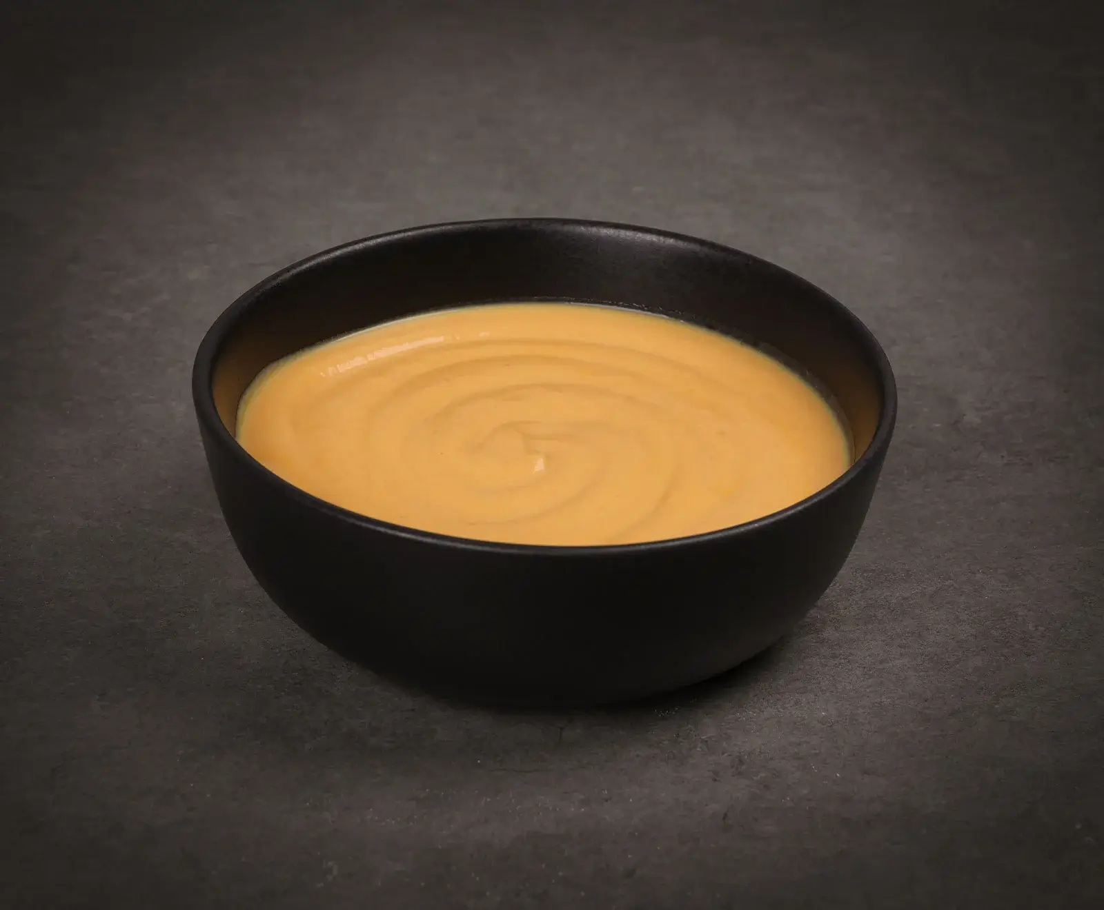 Eine schwarze Schale gefüllt mit einer glatten, orange-gelben Sauce oder Suppe, mit einem leichten Wirbelmuster auf der Oberfläche, auf einem dunklen, strukturierten Hintergrund.