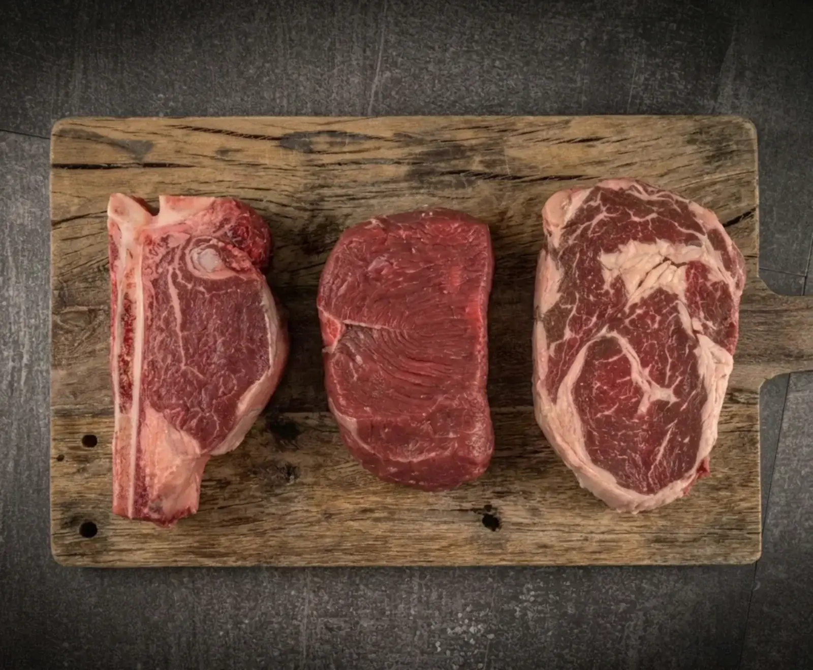 Drei rohe Rindersteaks – ein T-Bone, ein mageres Filet und ein marmoriertes Ribeye – liegen nebeneinander auf einem rustikalen Holzschneidebrett vor dunklem Hintergrund.