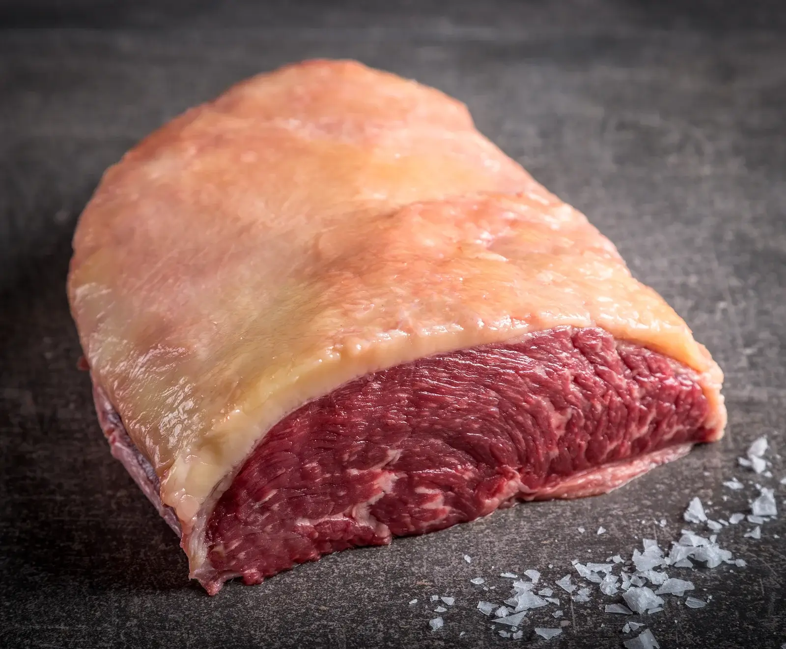Ein rohes Stück Rindfleisch, Picanha oder Tafelspitz, mit einer dicken, gelblichen Fettschicht oben und rotem Muskelfleisch unten, liegt auf einer dunklen Oberfläche mit groben Salzkristallen.
