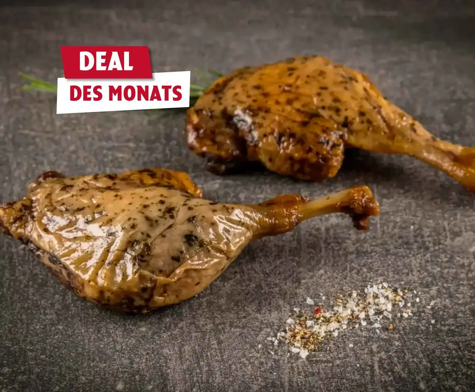 Zwei goldbraun gebratene Entenkeulen mit Kräutern auf dunklem Stein, daneben Gewürze und Rosmarin. Oben links ein Banner: "DEAL DES MONATS".