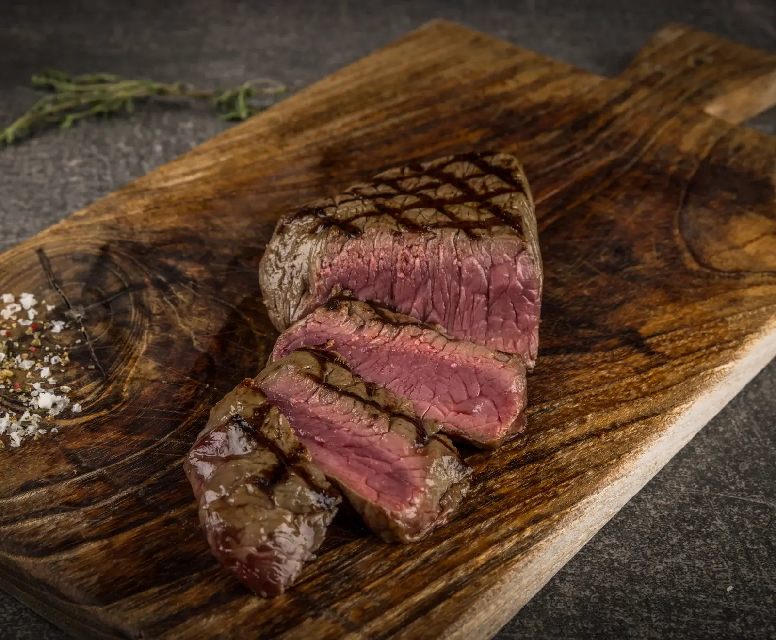 Ein saftiges, medium-rare gegrilltes Steak mit deutlichen Grillstreifen, aufgeschnitten auf einem rustikalen Holzbrett. Daneben liegen grobes Meersalz, Pfeffer und ein Kräuterzweig.