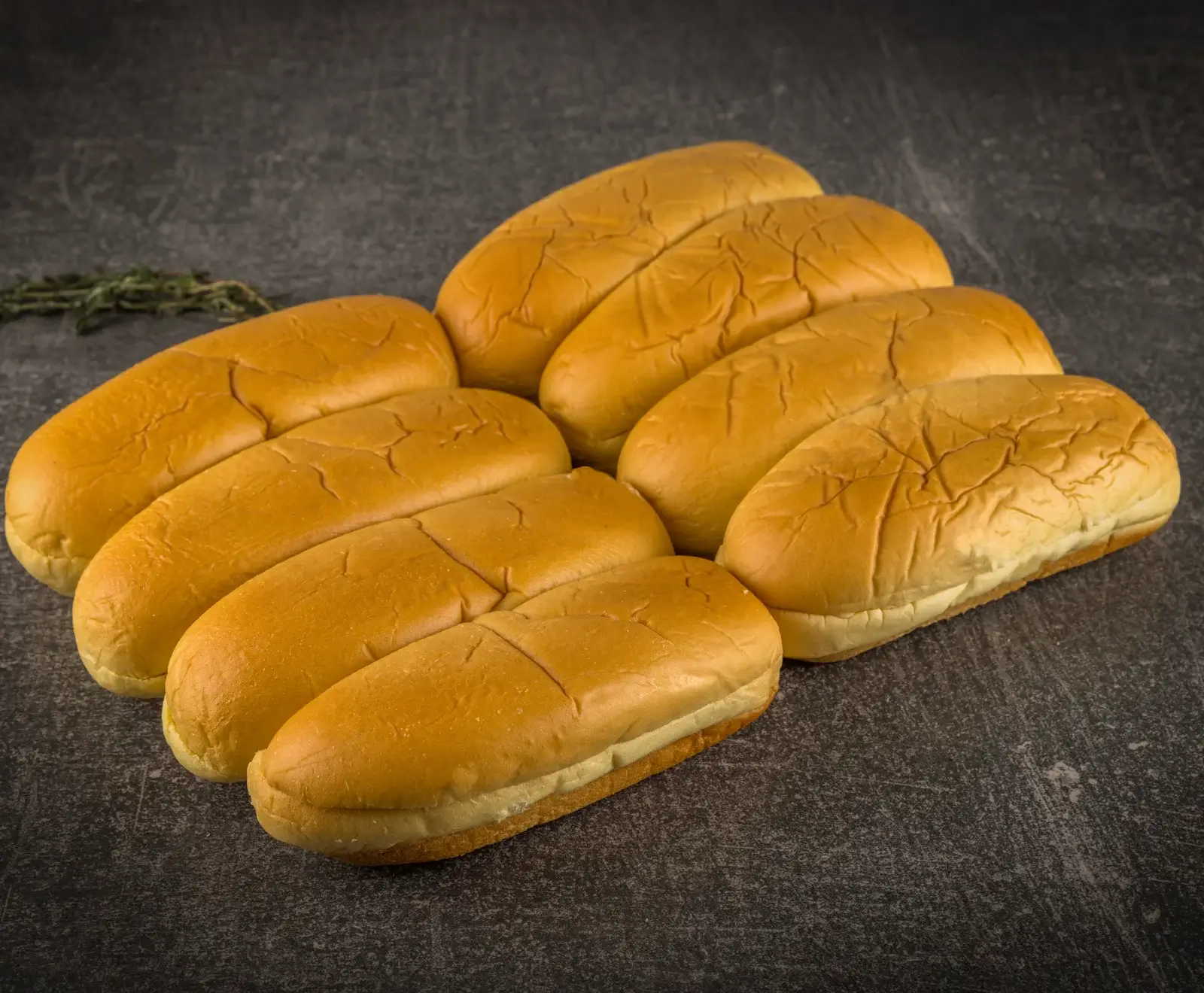 Mehrere goldbraune, weiche Hotdog-Brötchen liegen auf einer dunklen, strukturierten Oberfläche. Im Hintergrund ist ein grüner Kräuterzweig zu sehen.