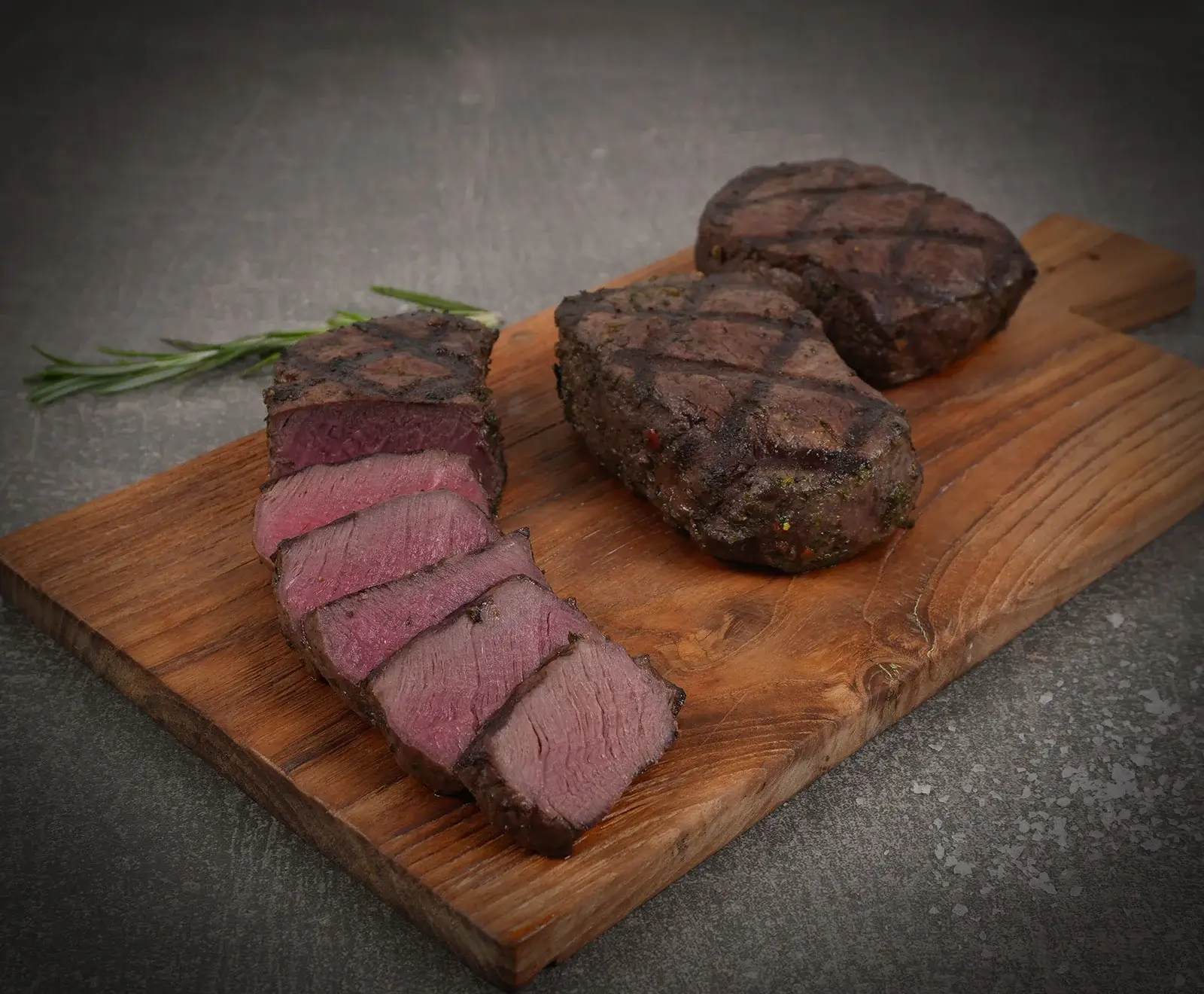 Drei gegrillte Steaks auf einem Holzschneidebrett. Eines ist in Scheiben geschnitten und zeigt einen rosa Kern. Zwei weitere Steaks liegen daneben. Ein Rosmarinzweig ist im Hintergrund zu sehen.