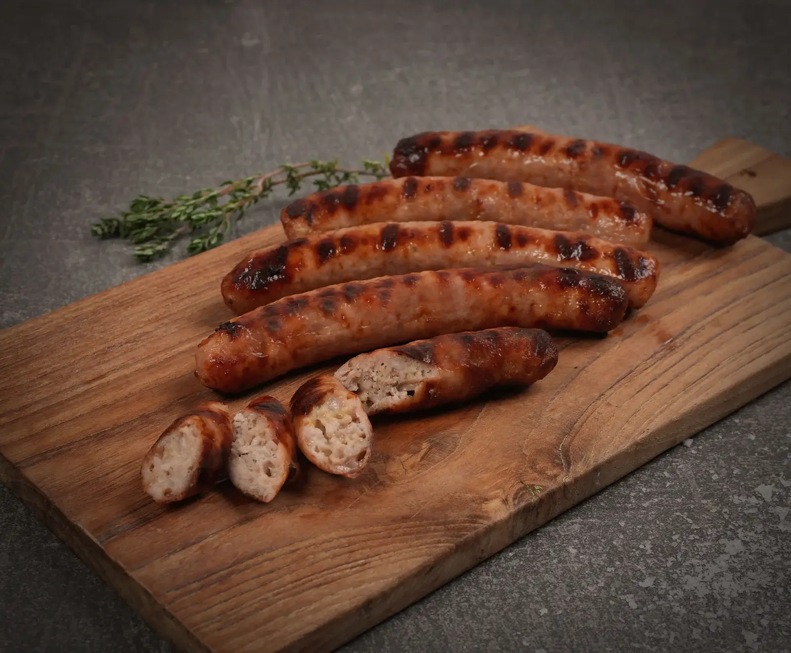 Fünf gegrillte Bratwürste auf einem Holzbrett, eine davon in Scheiben geschnitten, die das helle, körnige Innere zeigen. Ein Zweig Thymian liegt im Hintergrund auf einer dunklen Oberfläche.