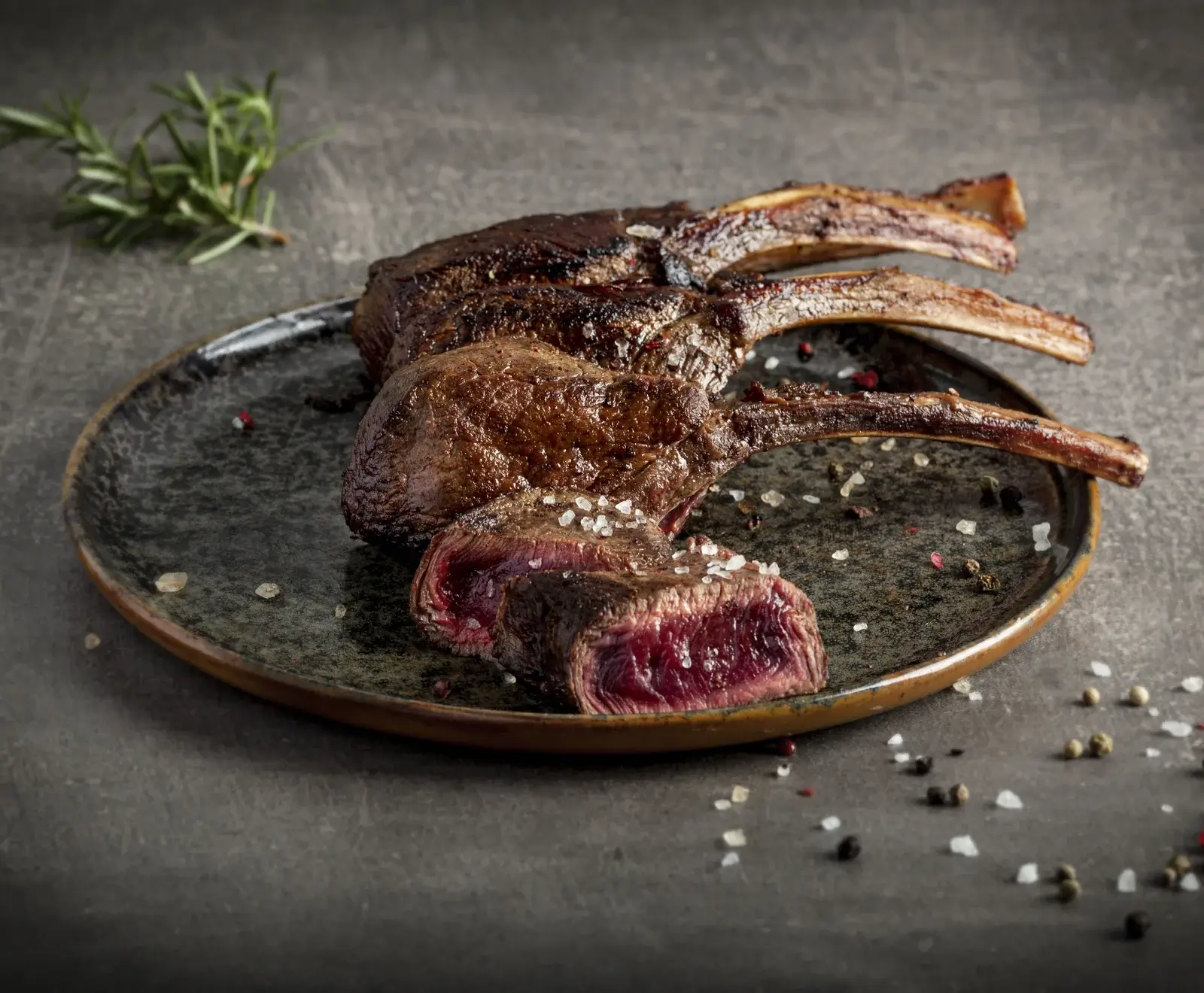 Drei gebratene Fleischkarrees mit Knochen auf einem dunklen Teller. Eines ist angeschnitten und zeigt eine medium-rare, rötliche Innenseite. Grobes Salz, Pfefferkörner und Rosmarin sind zu sehen.
