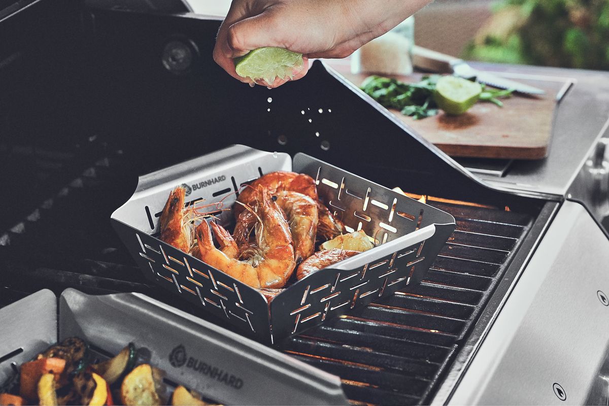 Eine Hand presst Limettensaft über gegrillte Garnelen in einem Burnhard-Grillkorb auf einem Gasgrill. Im Vordergrund sind Gemüse in einem weiteren Korb zu sehen, im Hintergrund eine Schneidebrett mit Limette und Kräutern.
