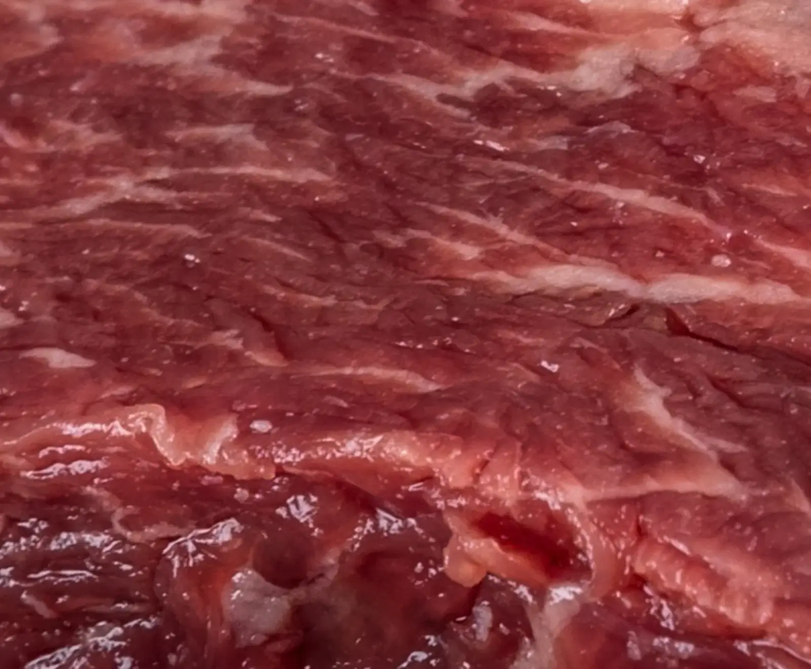 Nahaufnahme von rohem, dunkelrotem Fleisch mit deutlicher weißer Fettmarmorierung und glänzenden, feuchten Stellen.
