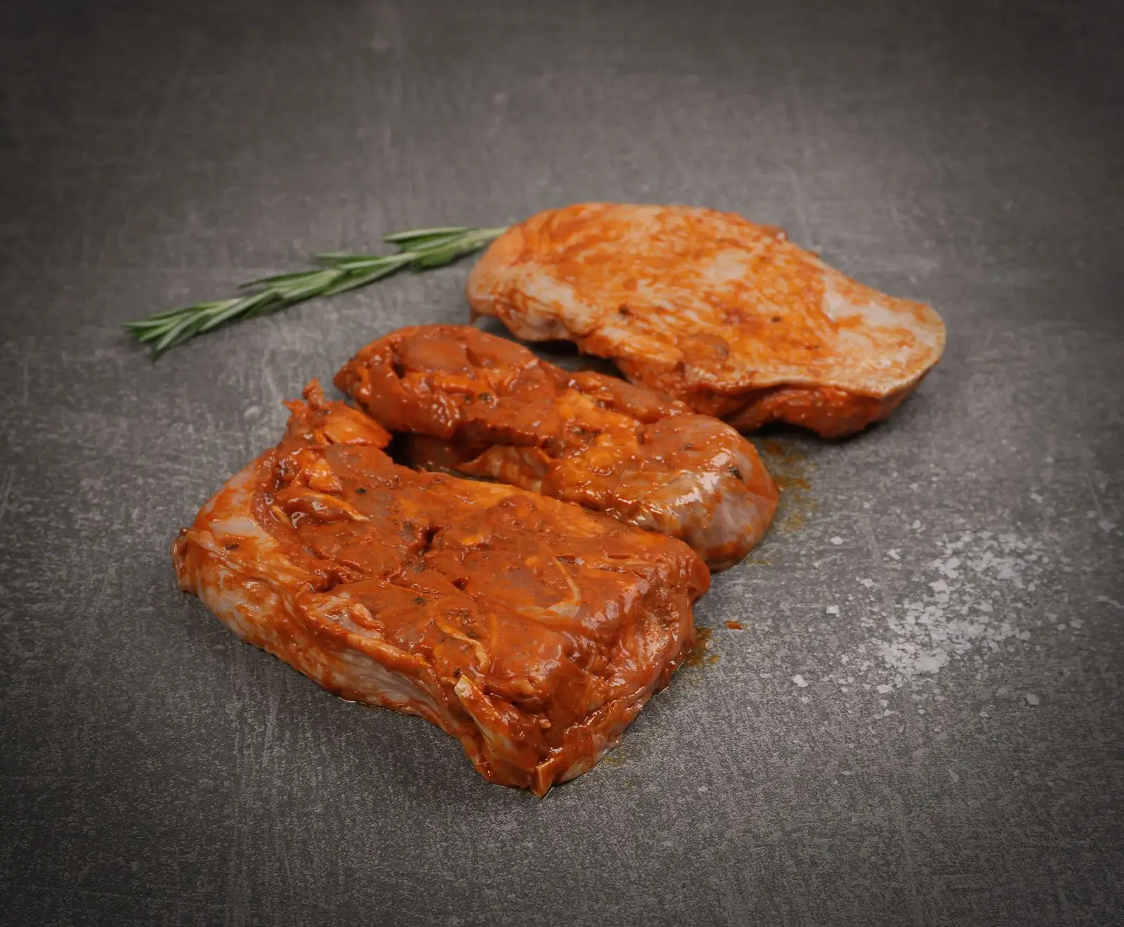 Drei rohe, rot marinierte Fleischstücke (z.B. Schweinehals oder Hähnchenbrust) mit einem Rosmarinzweig und grobem Salz auf dunklem, strukturiertem Untergrund.
