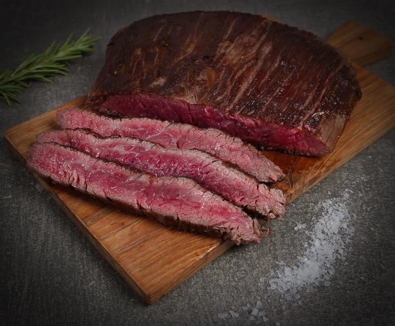 Ein aufgeschnittenes, medium-rare gegartes Steak auf einem Holzbrett, mit Rosmarin und grobem Meersalz auf dunklem Hintergrund.