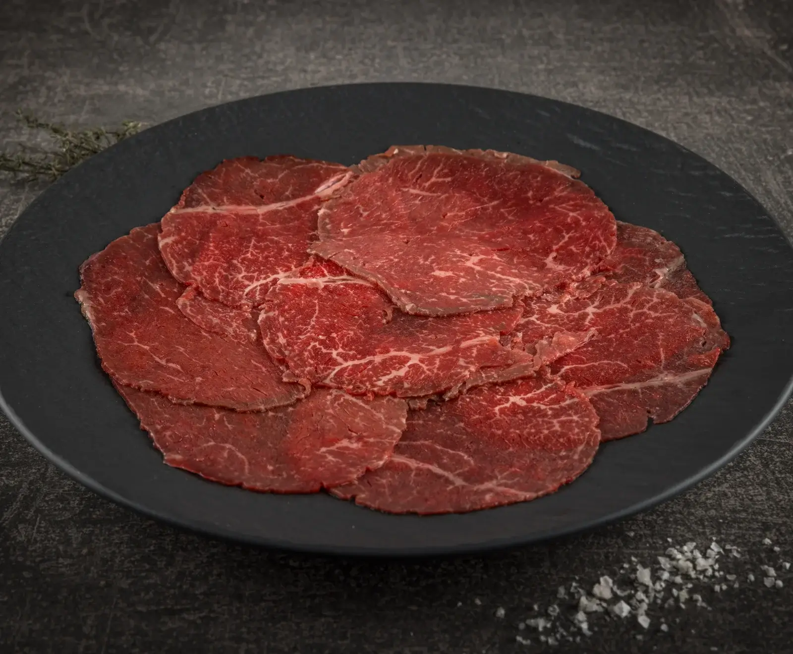 Dünn geschnittenes, rohes Rindfleisch mit feiner Marmorierung, auf einem dunklen Teller angerichtet. Im Hintergrund sind Kräuter und im Vordergrund grobes Salz erkennbar.