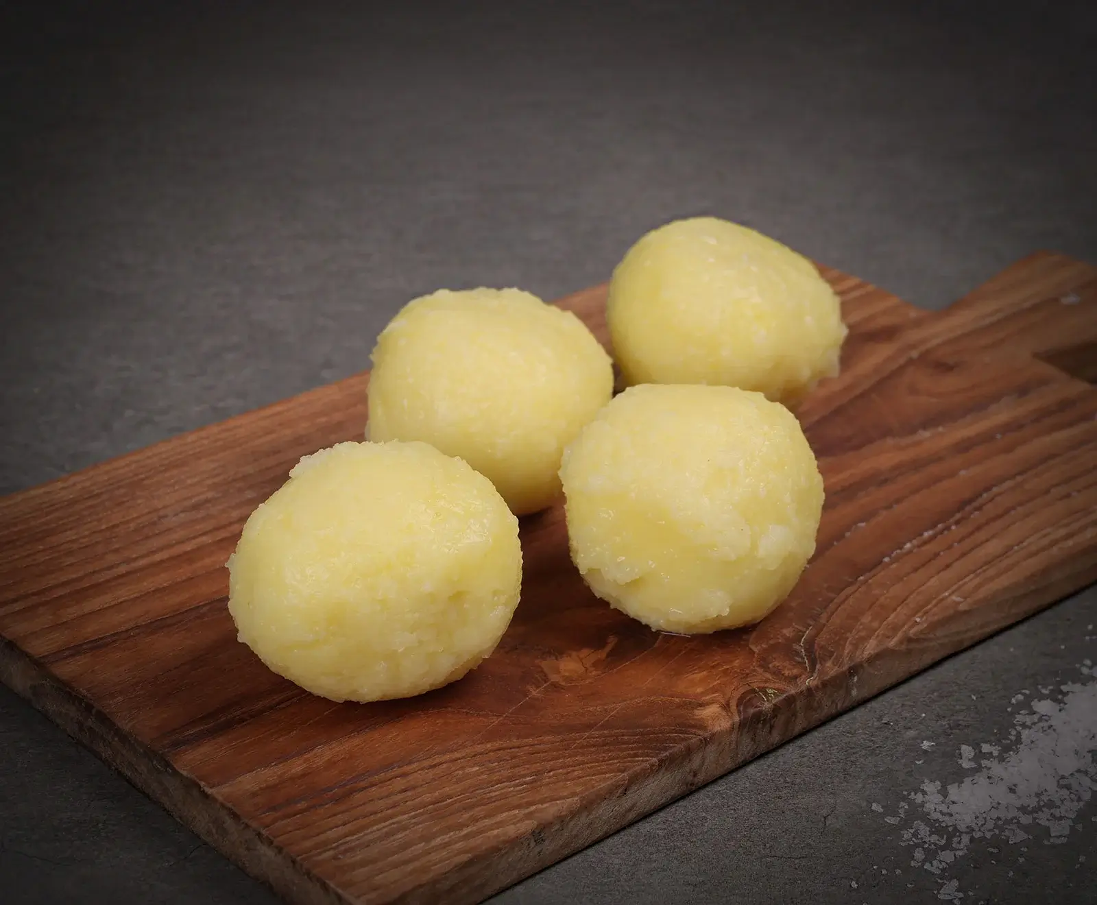 Vier goldgelbe, gekochte Kartoffelklöße liegen auf einem rustikalen Holzschneidebrett. Der dunkle Hintergrund zeigt leichte Salzkristalle.
