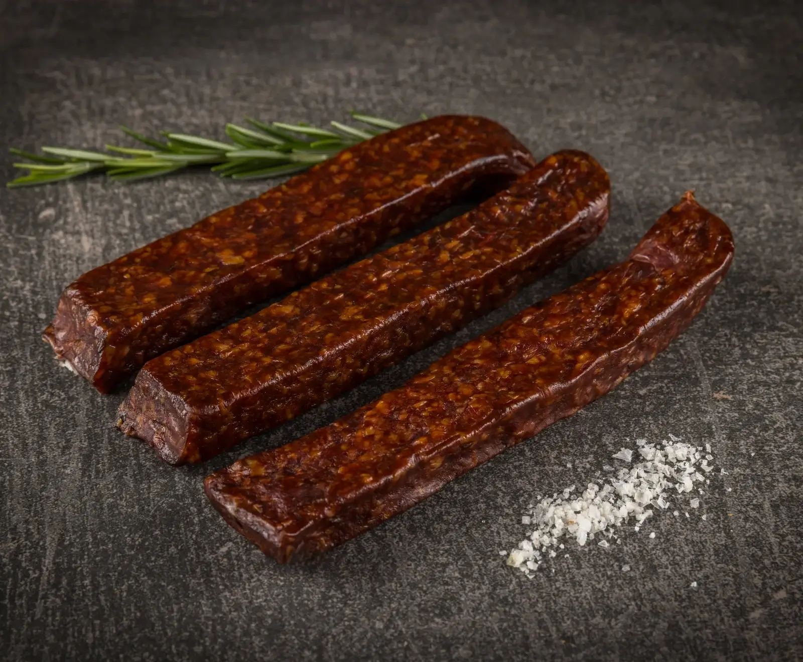 Drei dunkelbraune, rechteckige Wurststangen mit sichtbaren Fettstücken liegen diagonal auf einer dunklen Oberfläche, begleitet von einem Rosmarinzweig und grobem Meersalz.