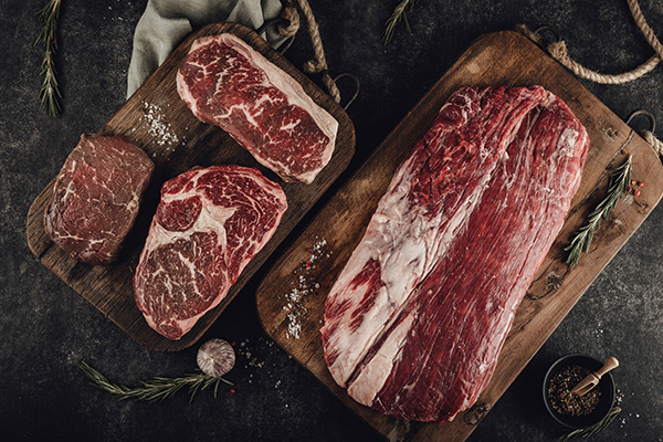 Verschiedene rohe Rindfleischstücke, darunter marmorierte Steaks und ein großes Flanksteak, liegen auf zwei rustikalen Holzbrettern. Grobes Salz, Rosmarin und Knoblauch sind auf dem dunklen Hintergrund verteilt.