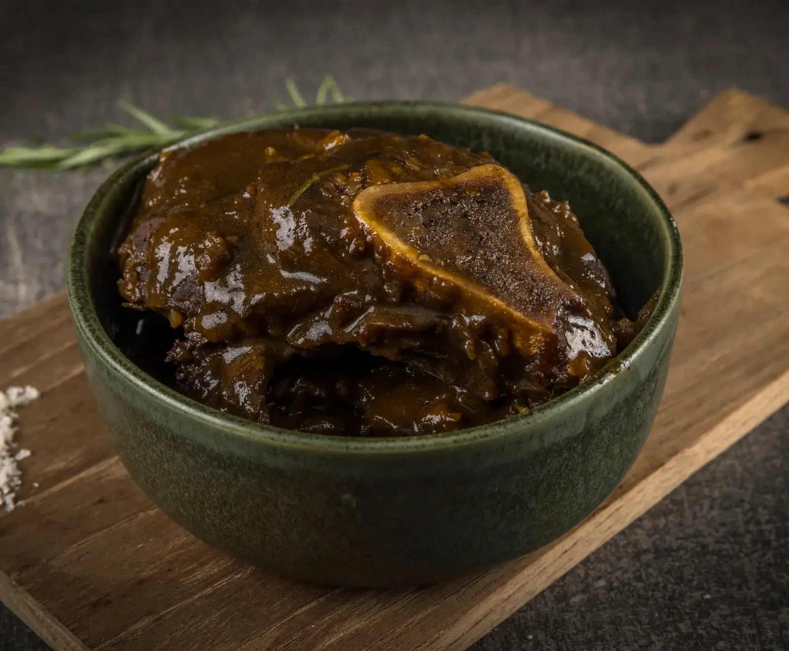Eine dunkelgrüne Schale gefüllt mit geschmortem Fleisch in dunkler, glänzender Soße, ein Knochenstück ist sichtbar. Die Schale steht auf einem Holzbrett, daneben ein Rosmarinzweig.