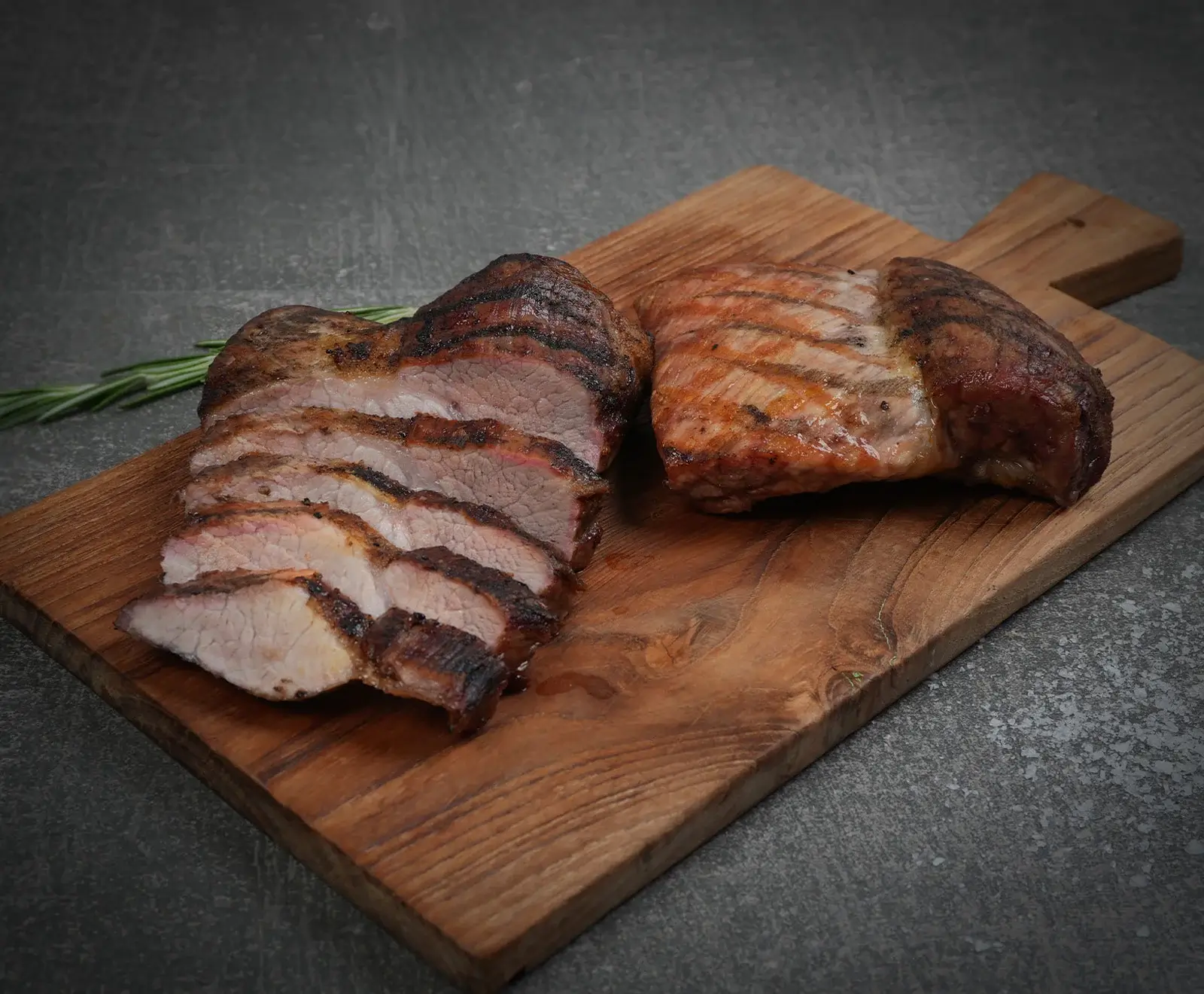 Zwei Stücke gegrilltes Fleisch auf einem Holzschneidebrett. Eines ist in Scheiben geschnitten und zeigt eine rosa Mitte, das andere ist ganz. Ein Rosmarinzweig liegt auf dem dunklen Hintergrund.