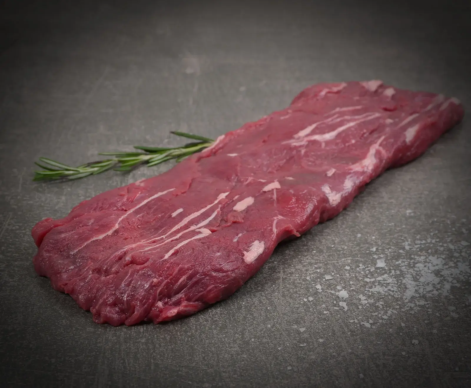 Ein rohes, flaches Rindersteak (Flank Steak) mit sichtbarer Marmorierung und zwei Rosmarinzweigen liegt auf einer dunklen, leicht gesprenkelten Oberfläche.