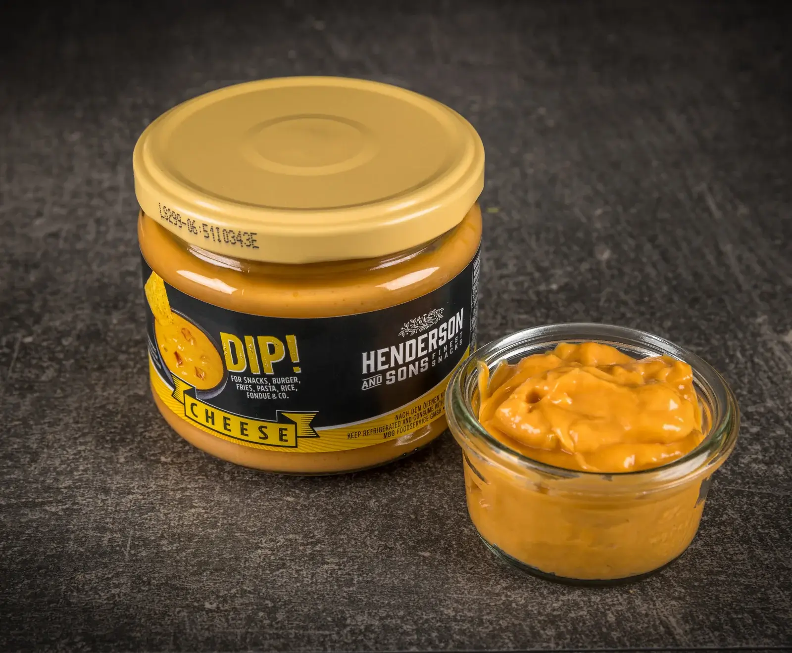 Ein Glas Henderson and Sons Käse-Dip mit gelbem Deckel und schwarzem Etikett steht neben einer kleinen Glasschale, die ebenfalls mit dem orangefarbenen Käse-Dip gefüllt ist, auf dunklem Untergrund.