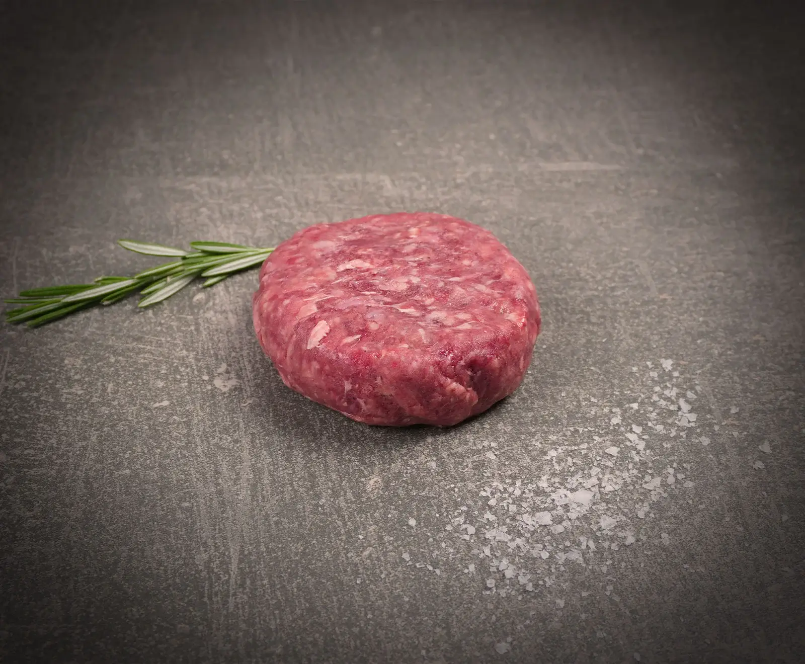 Ein rohes, rundes Burger-Patty liegt auf einer dunklen Steinoberfläche. Links daneben befindet sich ein Rosmarinzweig, rechts unten sind grobe Salzkristalle verstreut.