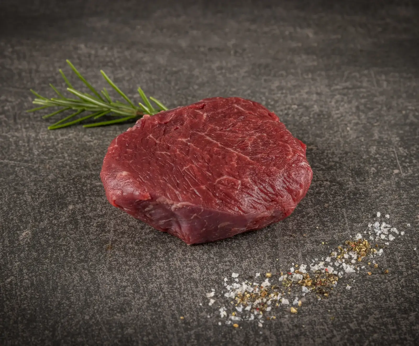 Ein rohes, rotes Rinderfilet liegt auf einer dunklen, strukturierten Oberfläche, daneben ein Rosmarinzweig und eine Prise grobes Salz und Pfeffer.