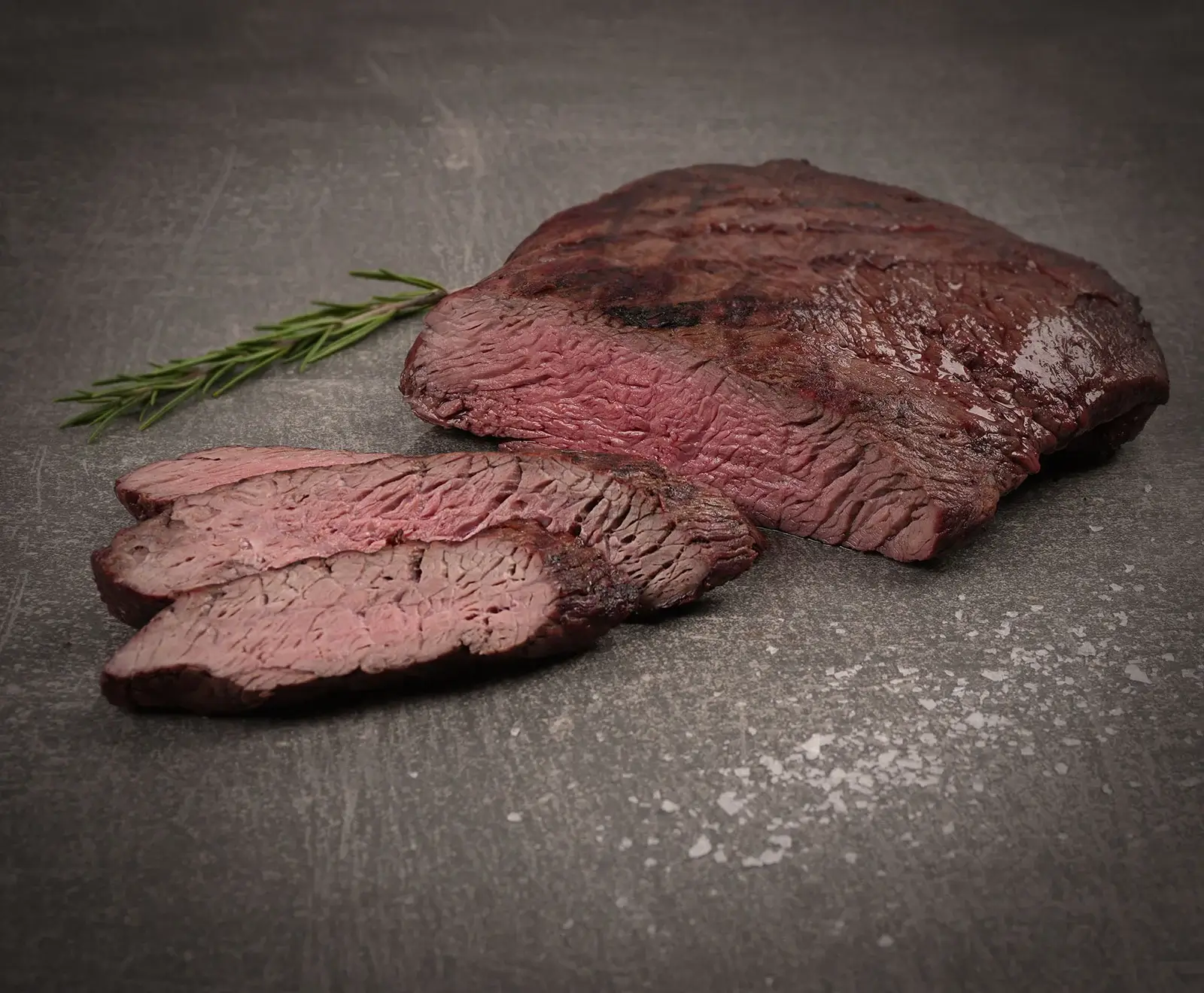 Ein aufgeschnittenes, medium-rosa gebratenes Steak mit Grillspuren, daneben ein Rosmarinzweig und grobes Salz auf dunklem Steinhintergrund.