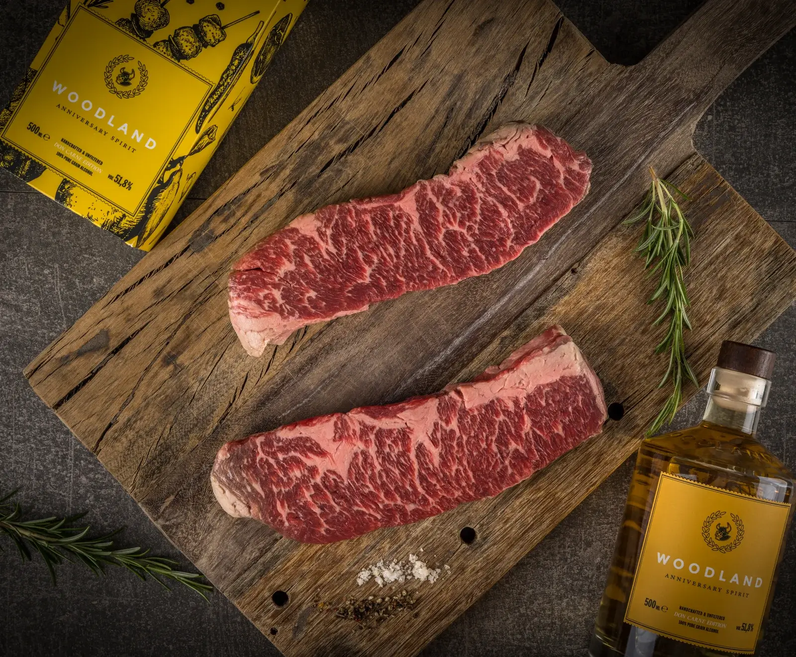 Zwei marmorierte Rohsteaks auf einem rustikalen Holzbrett, umgeben von Rosmarin, Salz, Pfeffer und einer Flasche sowie einer Box "Woodland Anniversary Spirit".