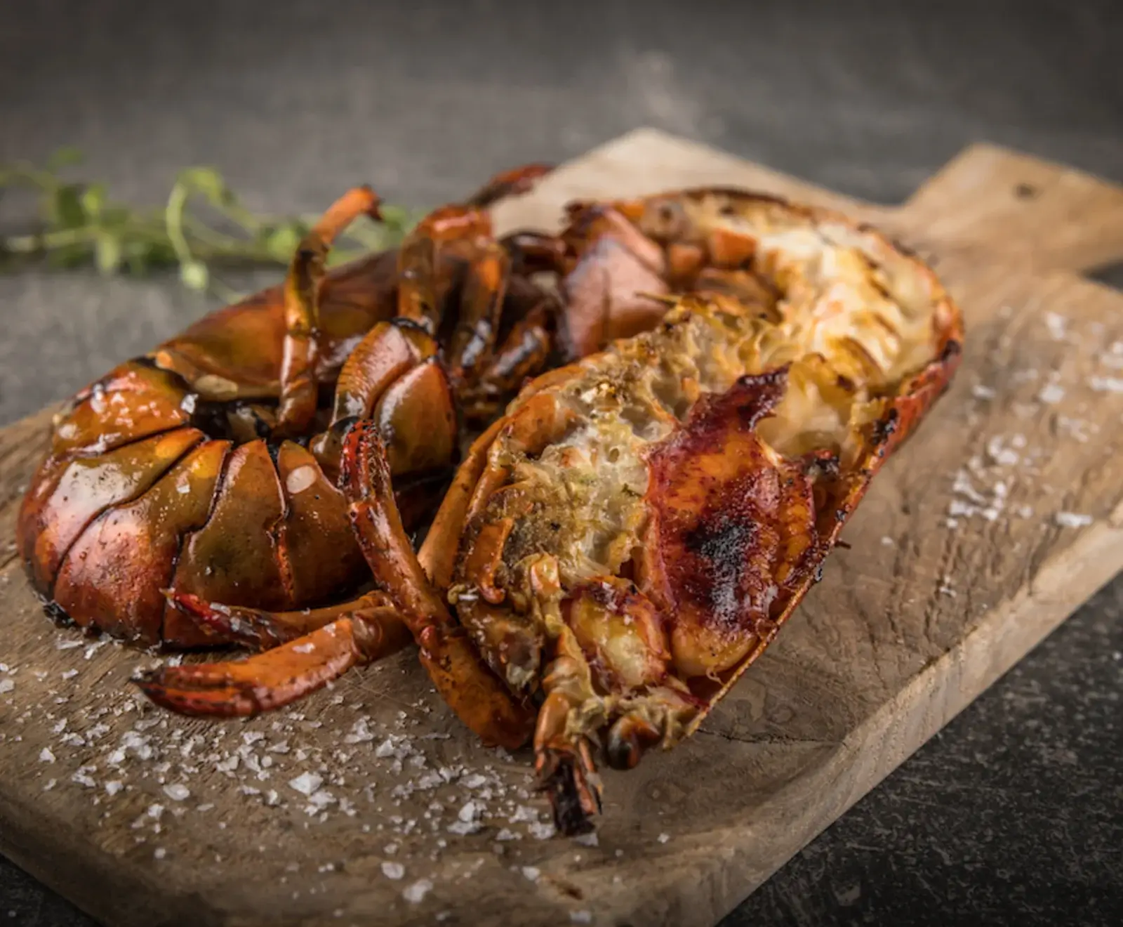 Ein halber, gegrillter Hummer mit goldbraunem Fleisch und roter Schale liegt auf einem rustikalen Holzbrett, bestreut mit grobem Meersalz. Ein Kräuterzweig ist im unscharfen Hintergrund zu sehen.