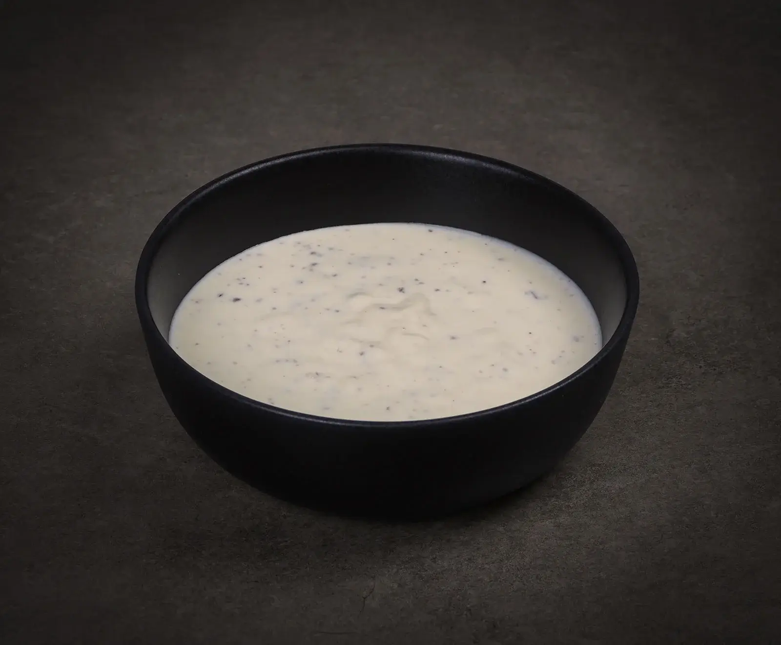 Eine schwarze Schale, gefüllt mit cremiger weißer Soße, die kleine dunkle Sprenkel (vermutlich Kräuter oder Pfeffer) enthält, auf einem dunklen, strukturierten Hintergrund.