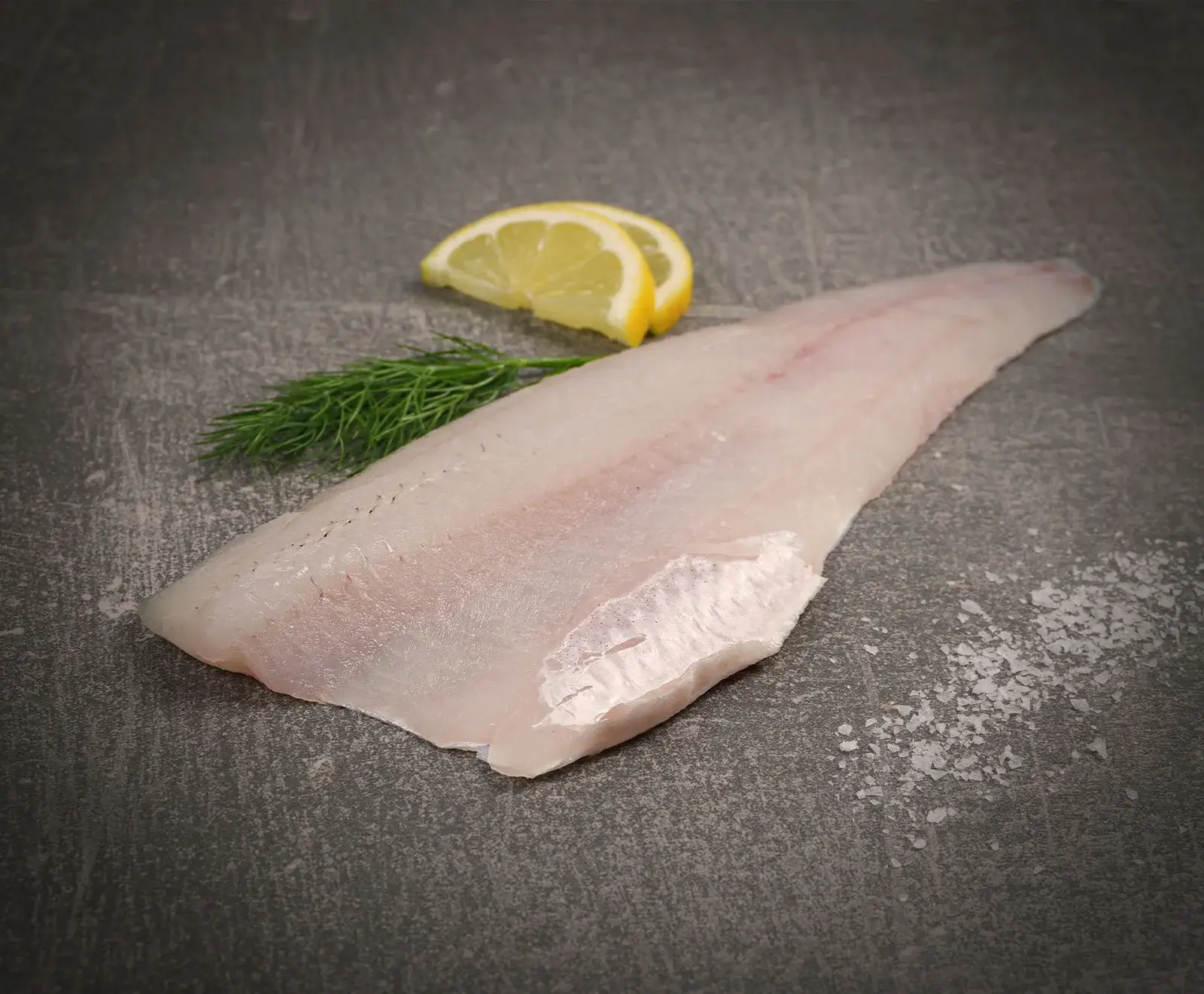 Ein rohes, helles Fischfilet liegt auf einer dunklen Oberfläche, daneben zwei Zitronenscheiben, ein Dillzweig und grobes Meersalz.