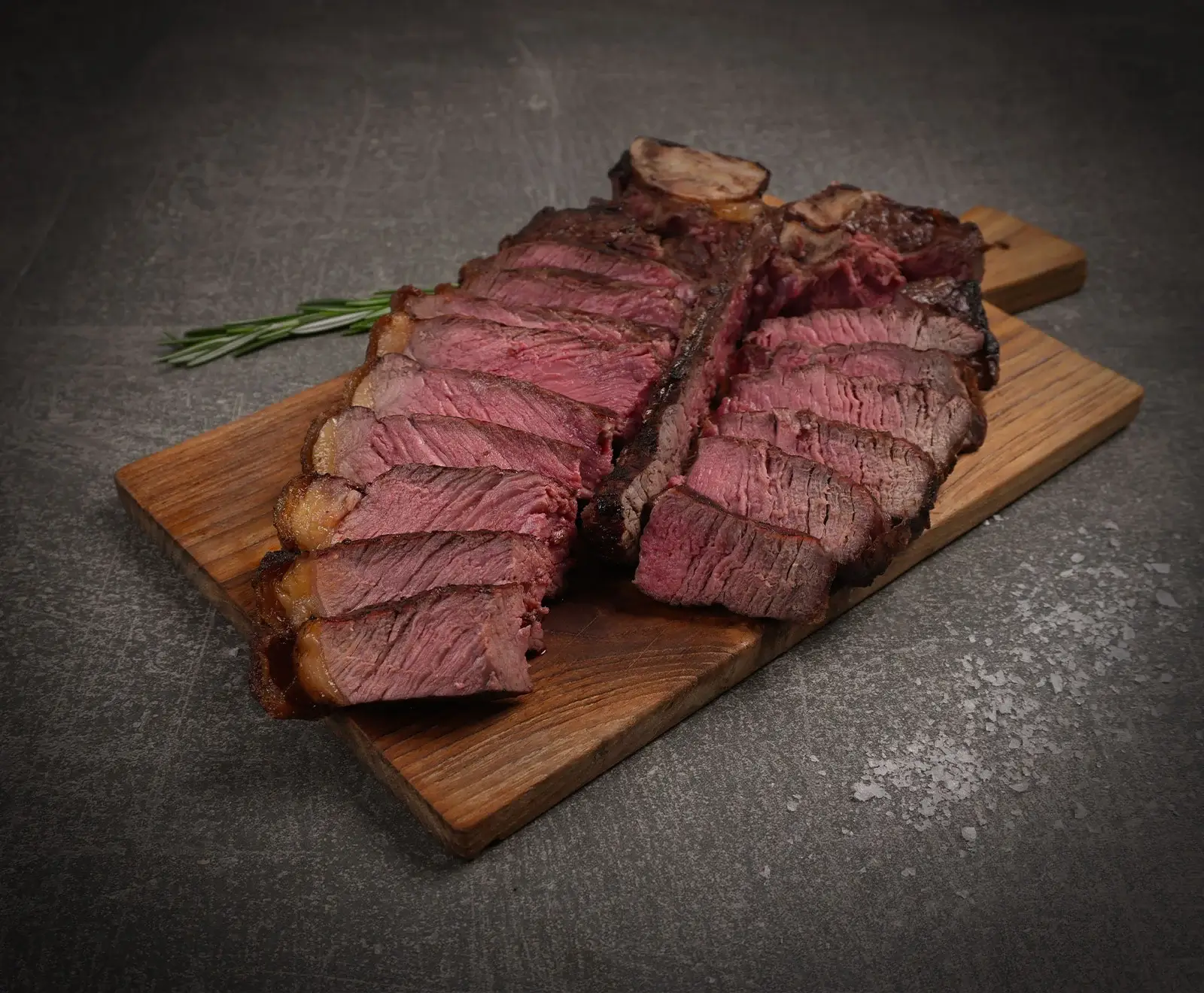 Ein in Scheiben geschnittenes, medium-rare T-Bone Steak auf einem Holzbrett. Daneben liegen ein Rosmarinzweig und grobes Salz auf einem dunklen Untergrund.