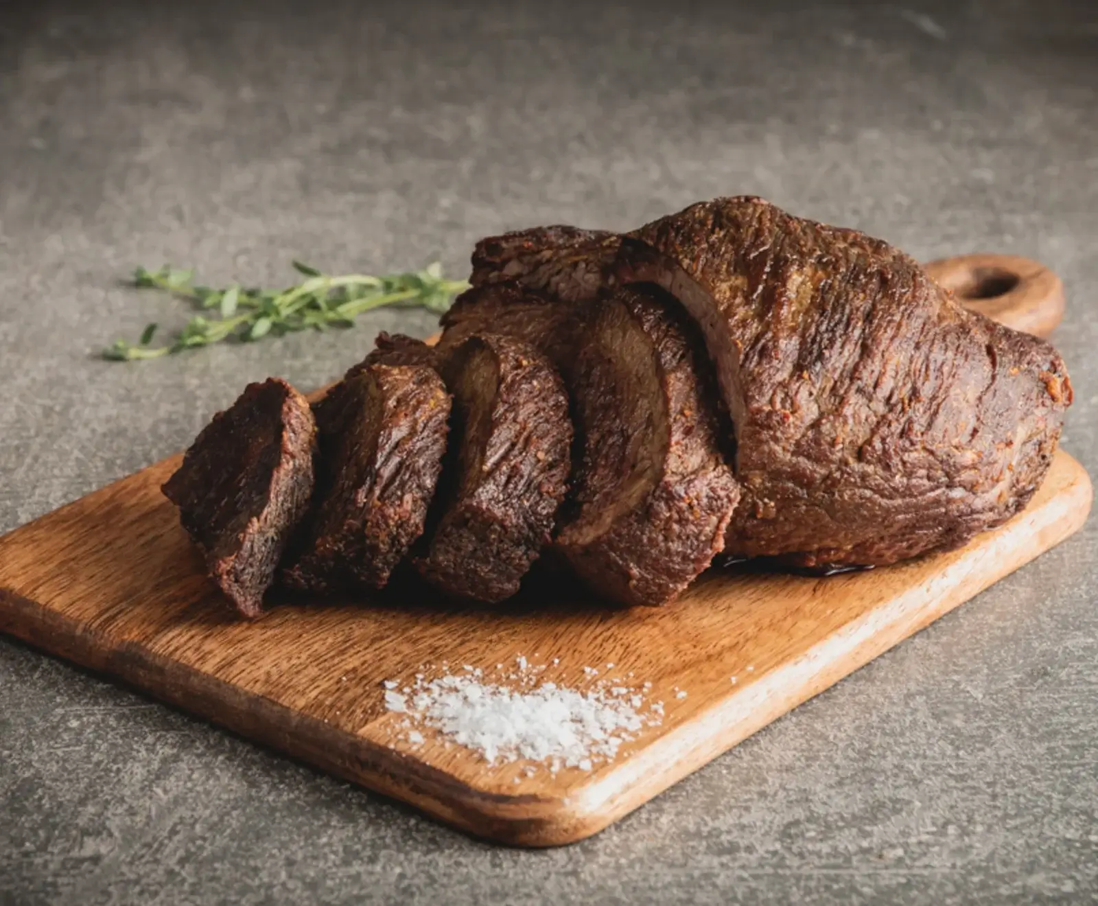 Ein großes, dunkelbraun gebratenes Stück Fleisch, teilweise in dicke Scheiben geschnitten, liegt auf einem Holzschneidebrett. Daneben sind grobes Meersalz und ein Zweig frischer Thymian zu sehen.