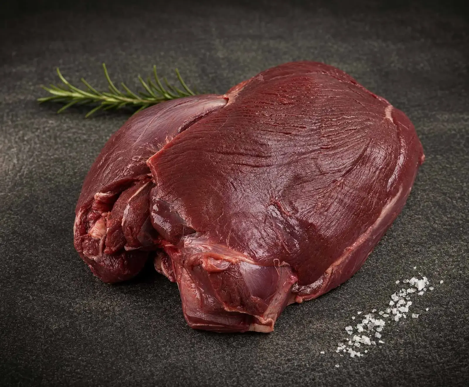 Ein großes Stück rohes, dunkelrotes Fleisch, vermutlich Wild oder Rind, liegt auf einer dunklen Oberfläche. Daneben sind ein Rosmarinzweig und grobes Meersalz zu sehen.