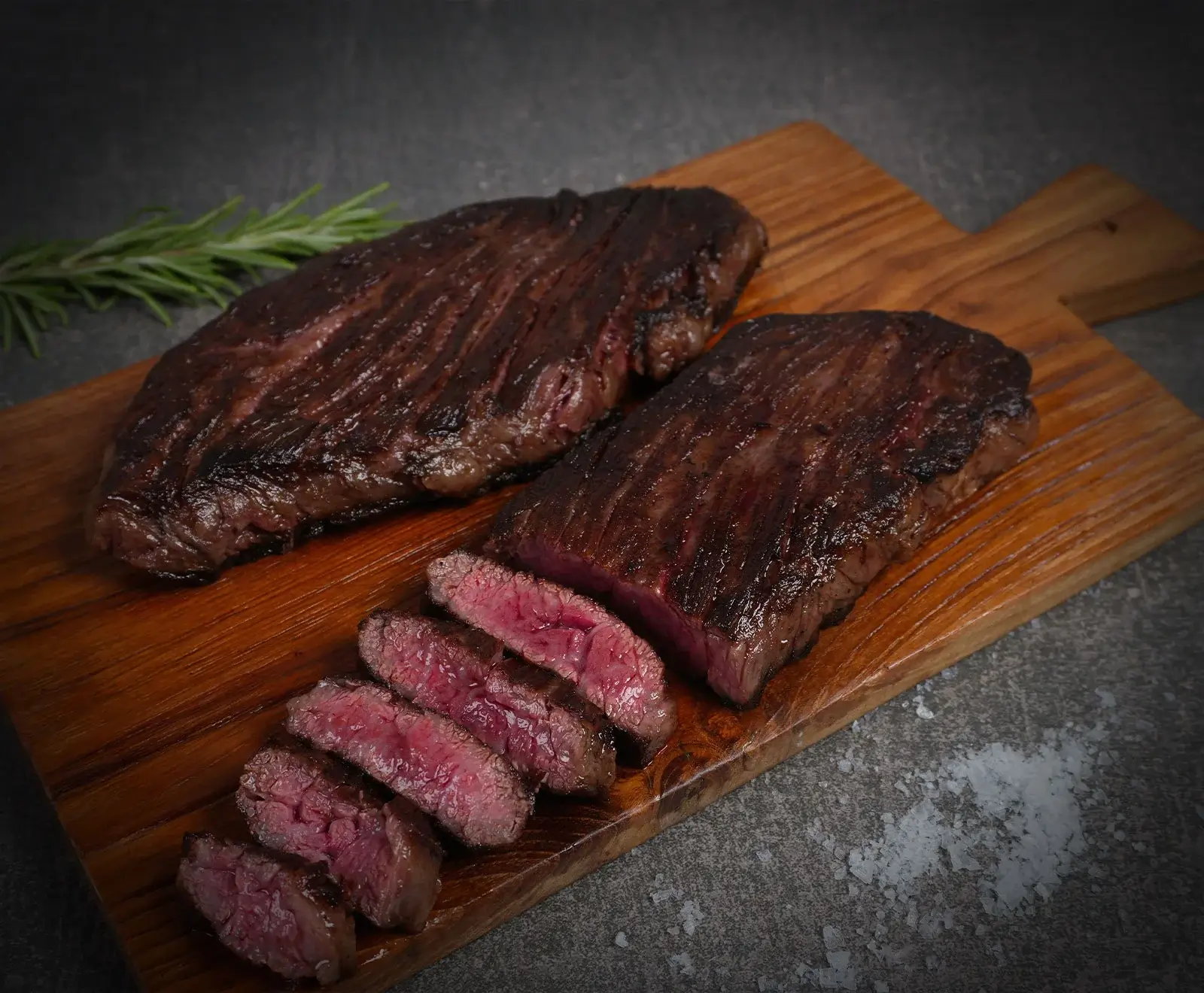 Zwei gegrillte Steaks auf einem Holzschneidebrett. Eines ist ganz, das andere in Scheiben geschnitten und zeigt eine rosa Mitte. Rosmarin und grobes Salz sind im Hintergrund zu sehen.