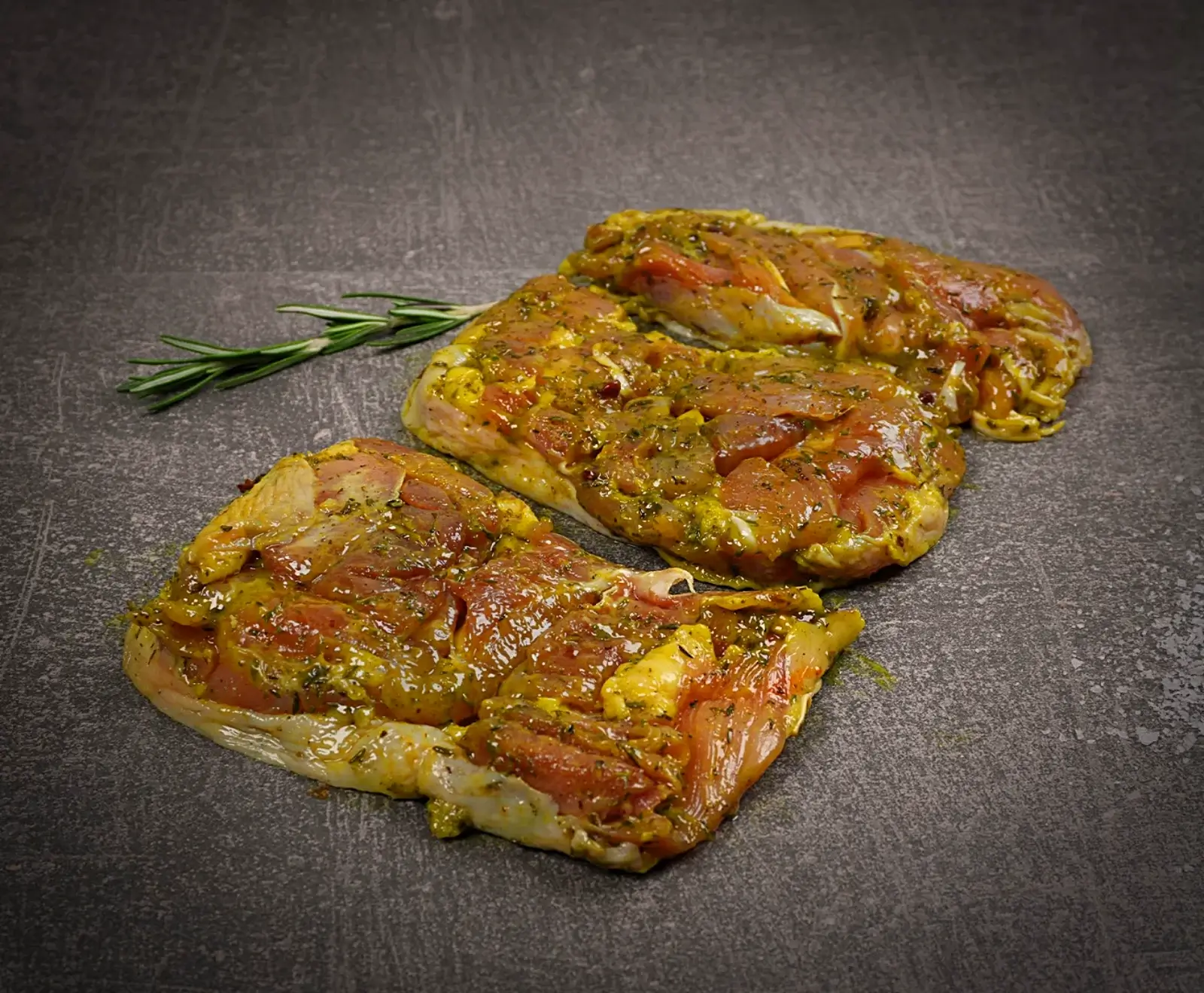 Drei rohe, marinierte Fleischstücke mit gelb-oranger Kräutermarinade liegen auf einem dunklen, strukturierten Untergrund. Daneben befindet sich ein frischer Rosmarinzweig.