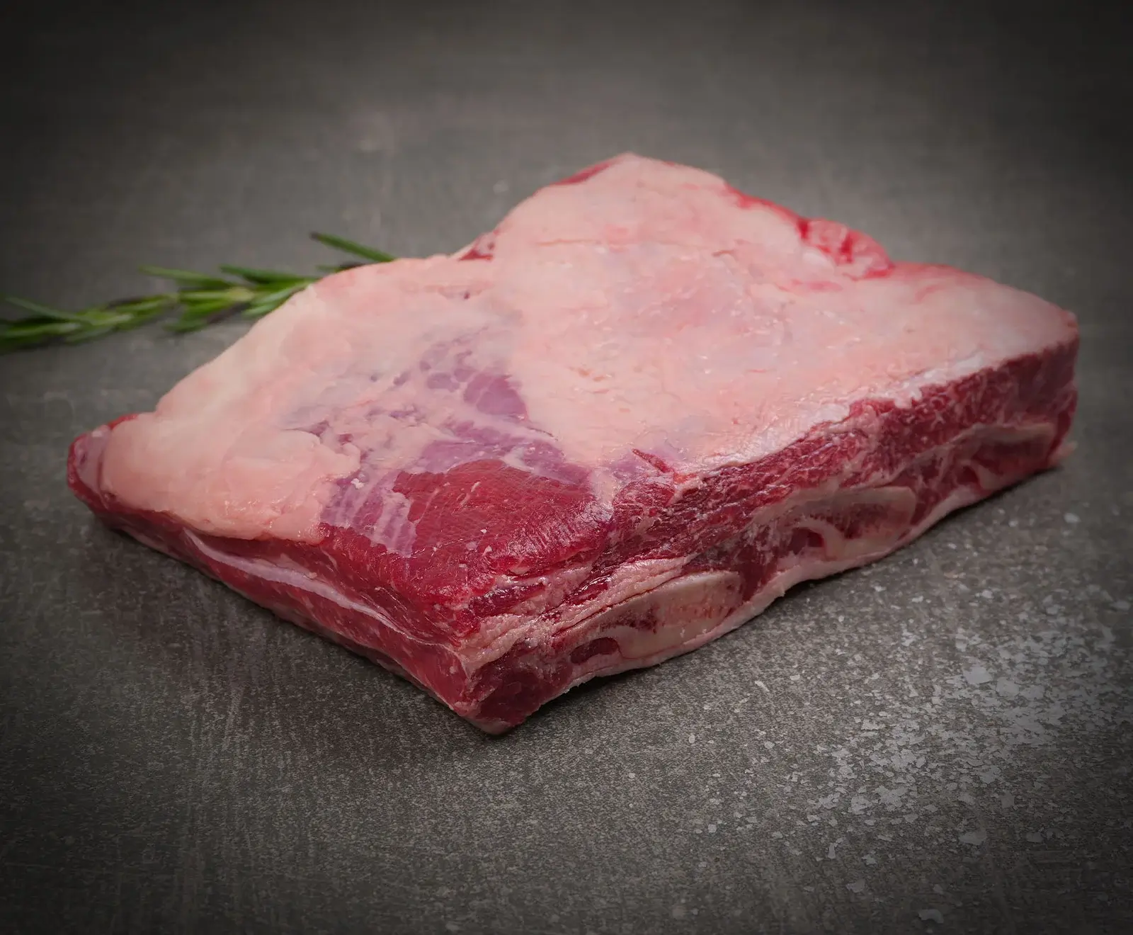 Ein großes, rohes Stück Rindfleisch mit einer dicken Fettschicht liegt auf einer dunklen, strukturierten Oberfläche. Links im Hintergrund ist ein Rosmarinzweig zu sehen.
