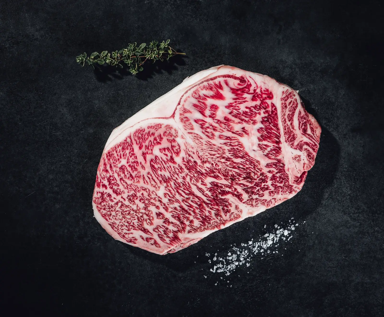 Ein rohes, stark marmoriertes Rindersteak liegt auf einer dunklen Oberfläche, begleitet von einem Thymianzweig und grobem Meersalz.