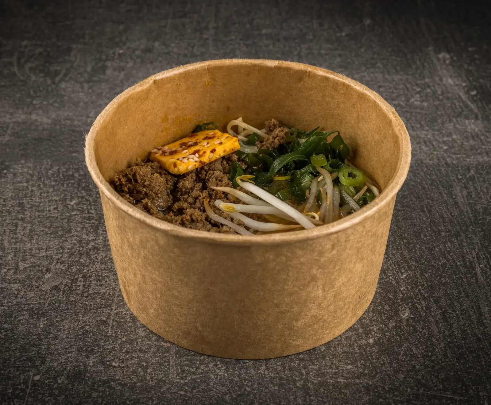 Ein brauner Pappbecher mit asiatischem Nudelgericht: Hackfleisch, Nudeln, Sojasprossen, Frühlingszwiebeln und ein gewürzter Tofublock mit Sesam. Auf dunklem Hintergrund.