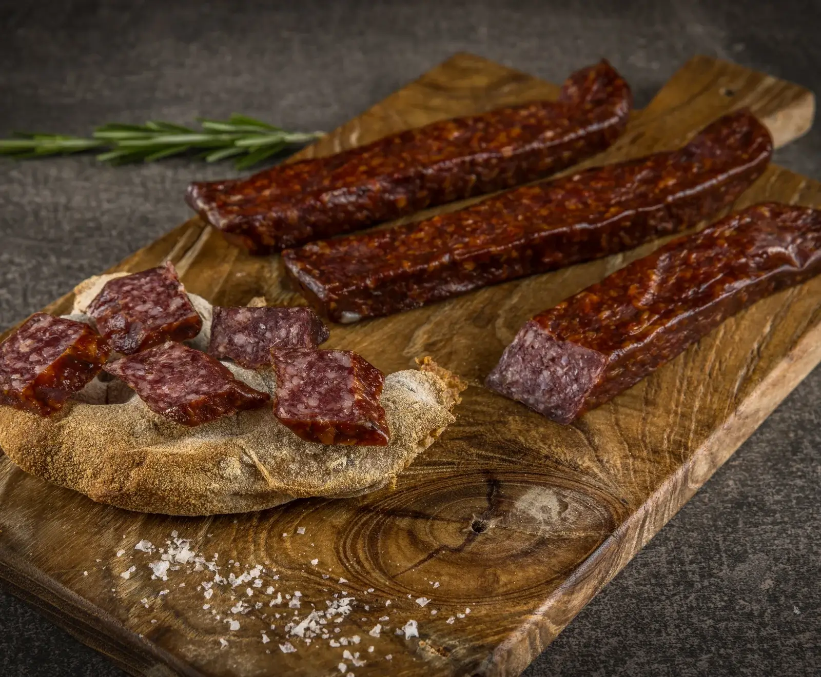 Drei dunkle, rechteckige Salami- oder Rohwurststücke auf einem Holzschneidebrett. Eines ist angeschnitten, mit Würfeln auf rustikalem Brot. Rosmarin und grobes Salz sind ebenfalls zu sehen.
