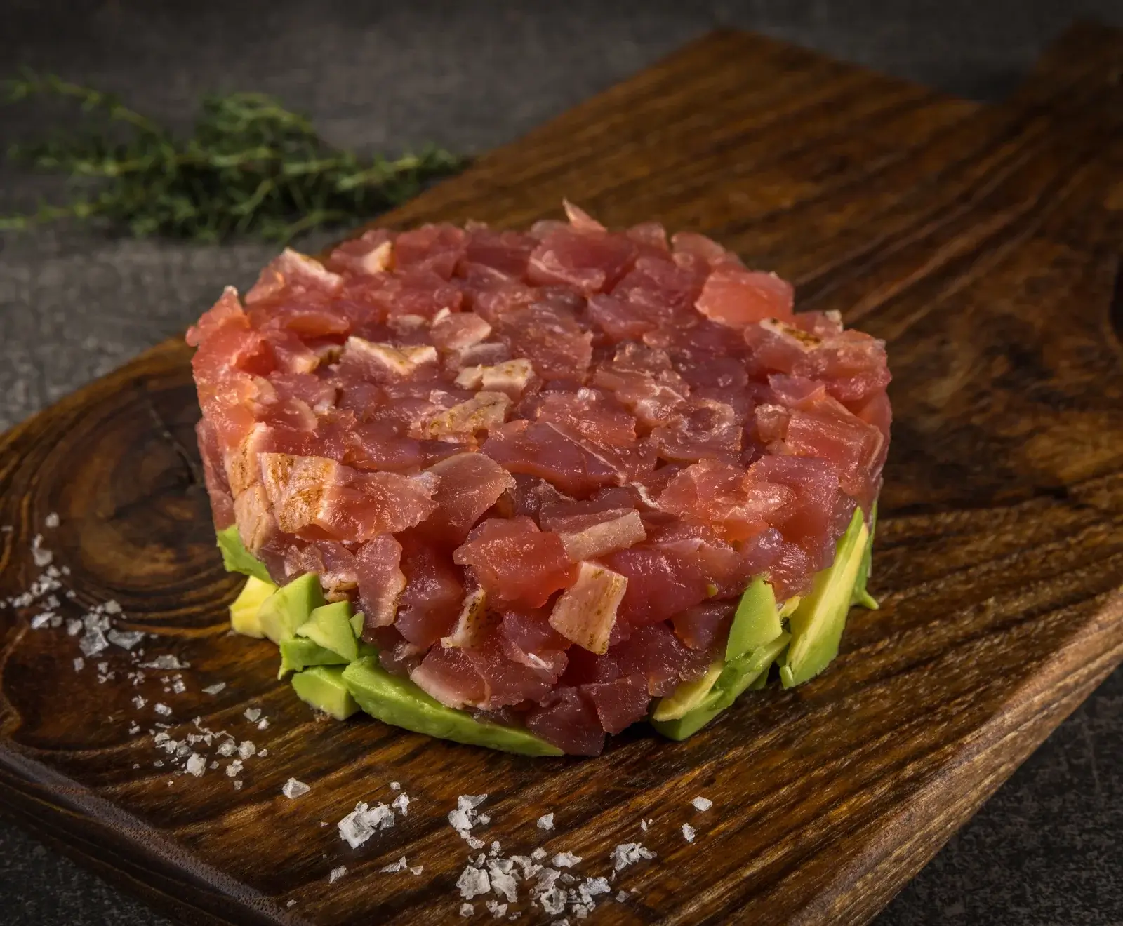 Ein rundes Thunfisch-Tatar mit gewürfeltem Avocado als Basis, serviert auf einem dunklen Holzbrett mit grobem Meersalz. Im Hintergrund sind Kräuter zu sehen.
