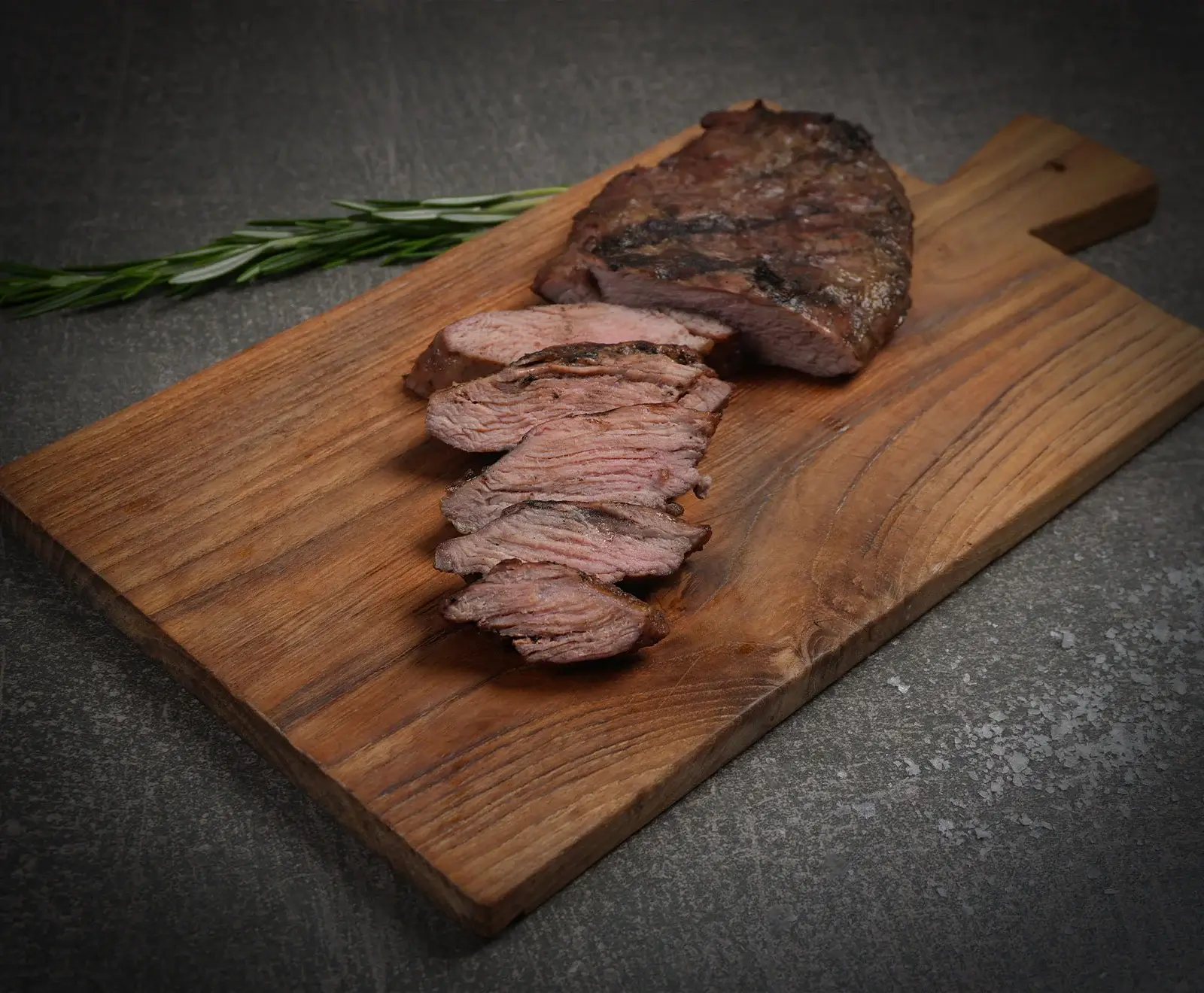 Ein saftiges, medium-rare gegrilltes Steak, teilweise in Scheiben geschnitten, liegt auf einem rustikalen Holzschneidebrett. Ein frischer Rosmarinzweig ist daneben auf dunklem Hintergrund platziert.