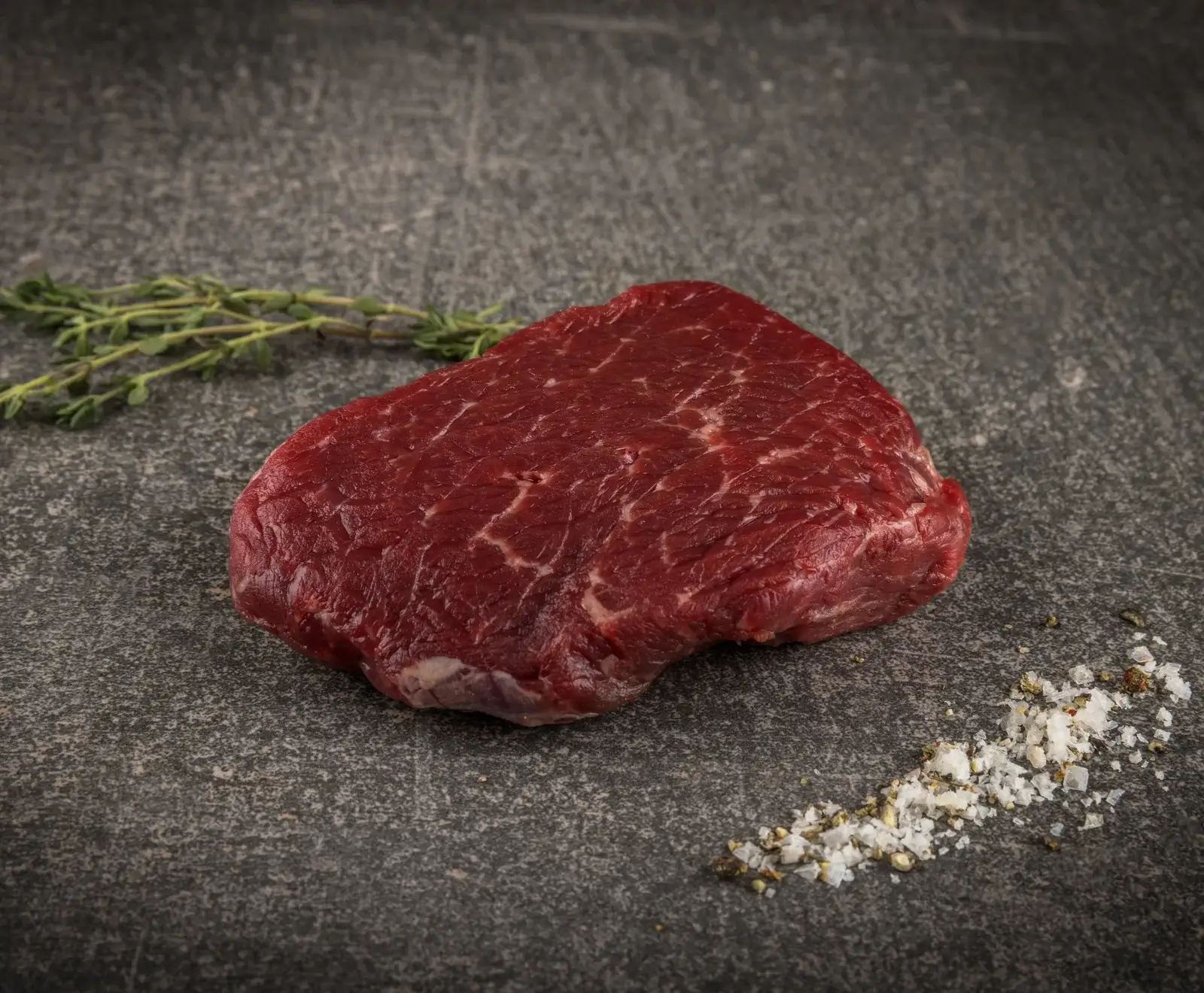Ein rohes, rotes Stück Rindfleisch (Steak) liegt auf einer dunklen Steinoberfläche, daneben frischer Thymian und eine Prise grobes Salz mit Pfeffer.