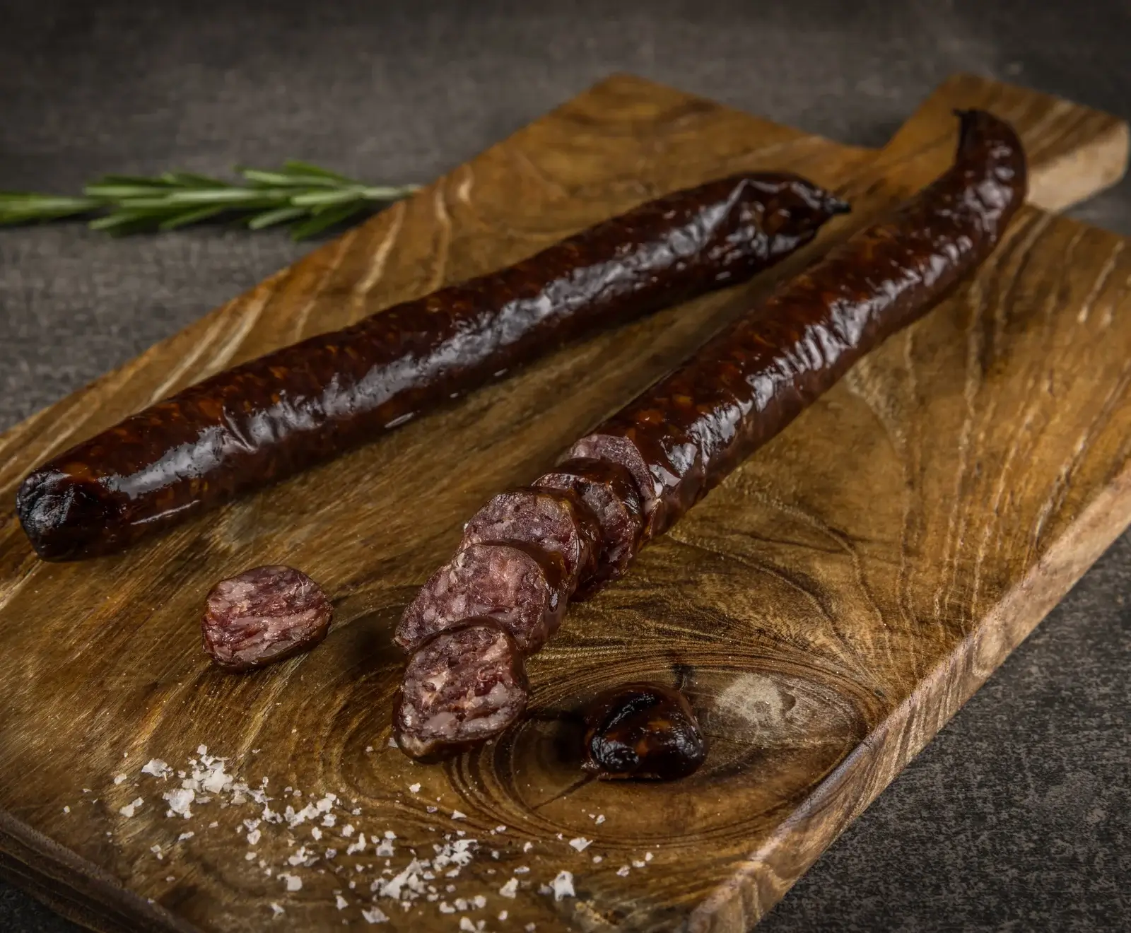 Zwei dunkle, glänzende Rohwürste auf einem Holzbrett, eine davon in Scheiben geschnitten, mit grobem Salz und einem Rosmarinzweig im Hintergrund auf dunklem Untergrund.