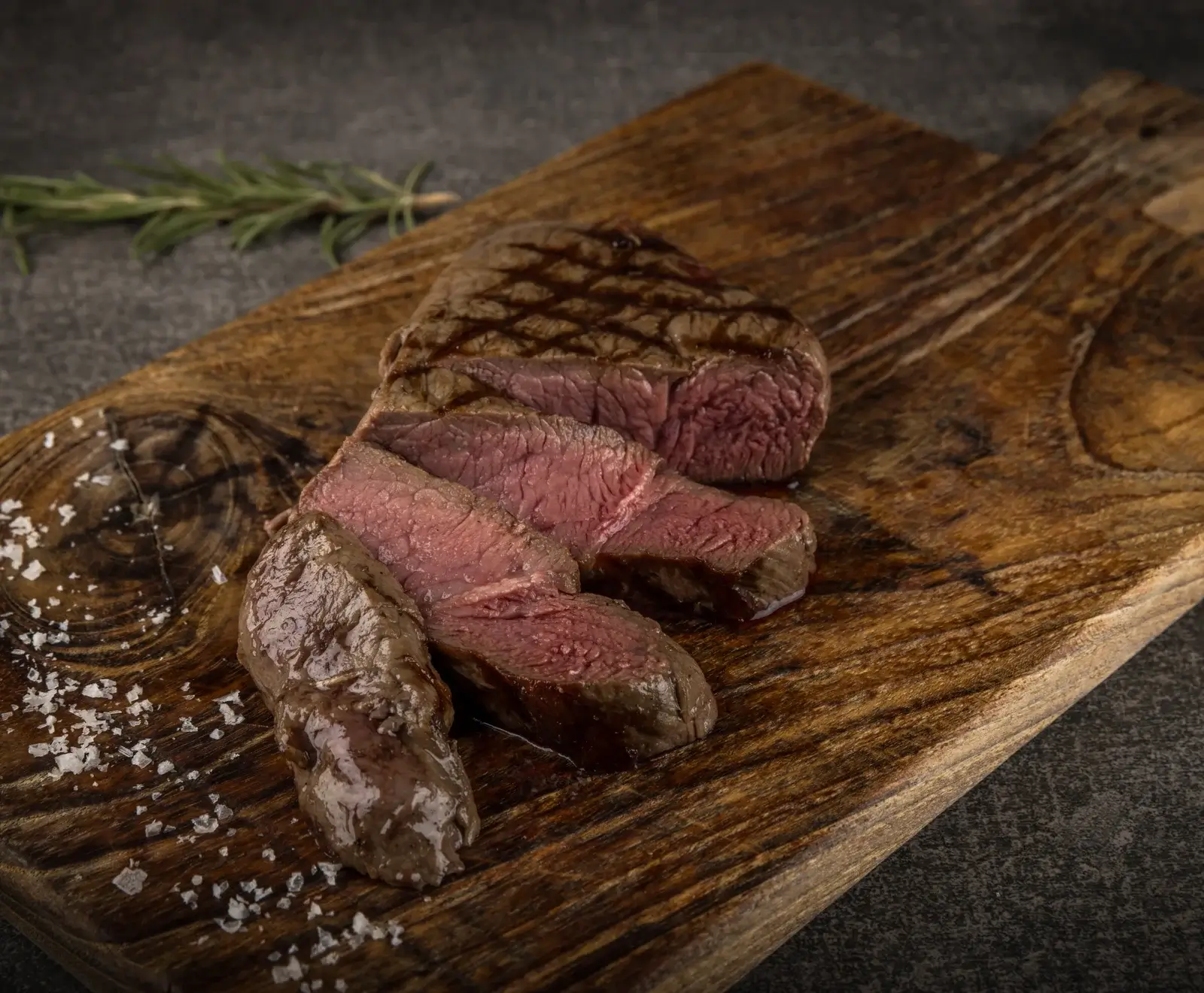 Ein perfekt gegrilltes, in Scheiben geschnittenes Steak, medium-rare, mit Grillstreifen, auf einem rustikalen Holzbrett. Grobes Meersalz und ein Rosmarinzweig sind im Hintergrund zu sehen.