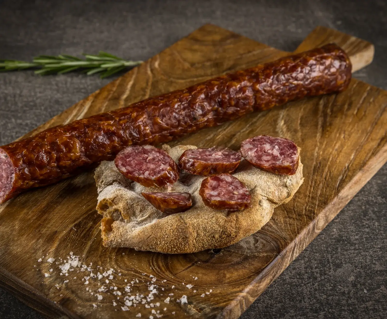 Eine lange, dunkelrote Rohwurst liegt auf einem Holzschneidebrett. Daneben sind Scheiben der Wurst auf rustikalem Brot angerichtet, mit grobem Salz und einem Rosmarinzweig im Hintergrund.