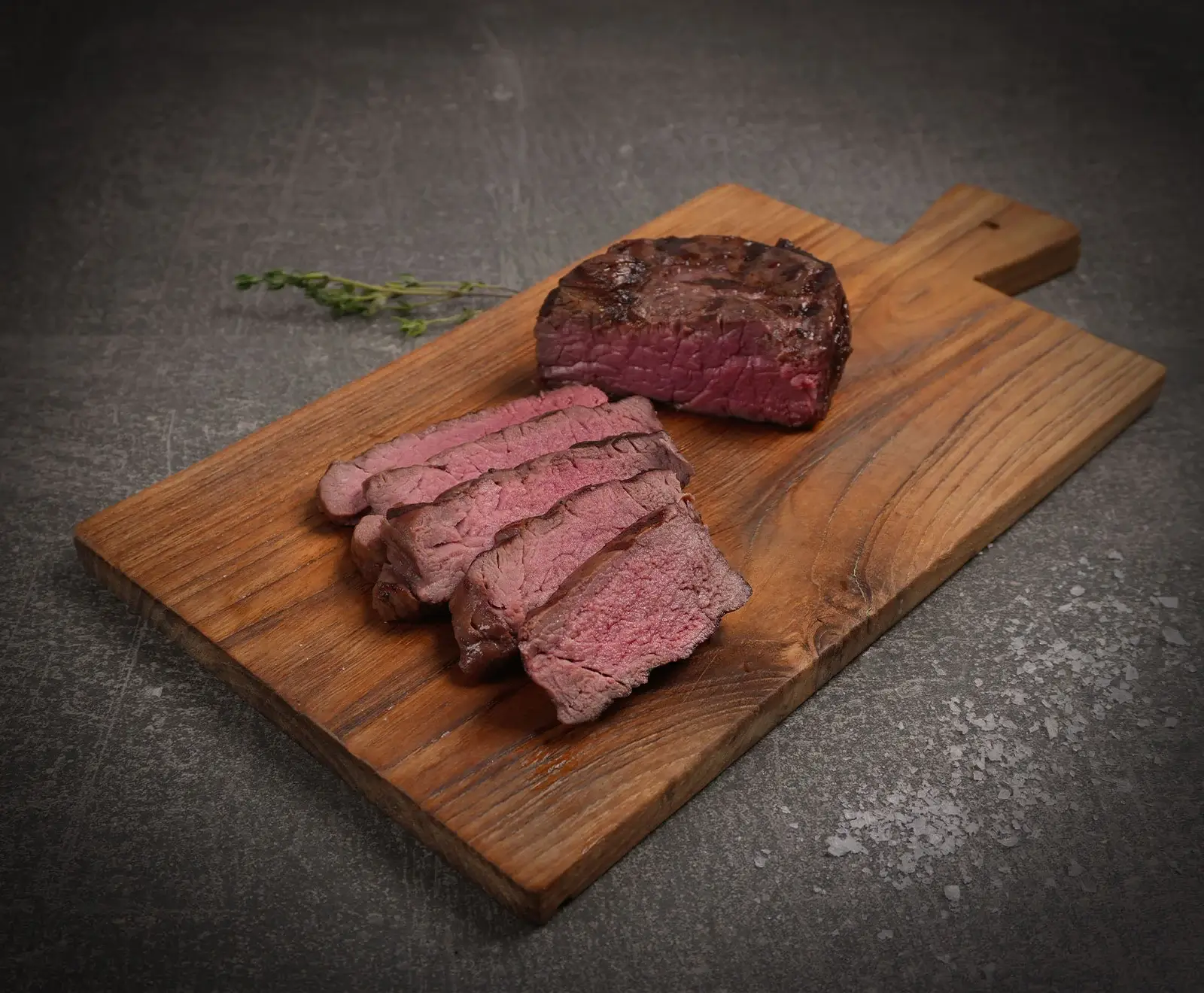 Ein perfekt gegartes, medium-rare Rindersteak, teils geschnitten, teils ganz, liegt auf einem Holzschneidebrett. Dazu ein Zweig Thymian und Salzflocken auf dunklem Untergrund.