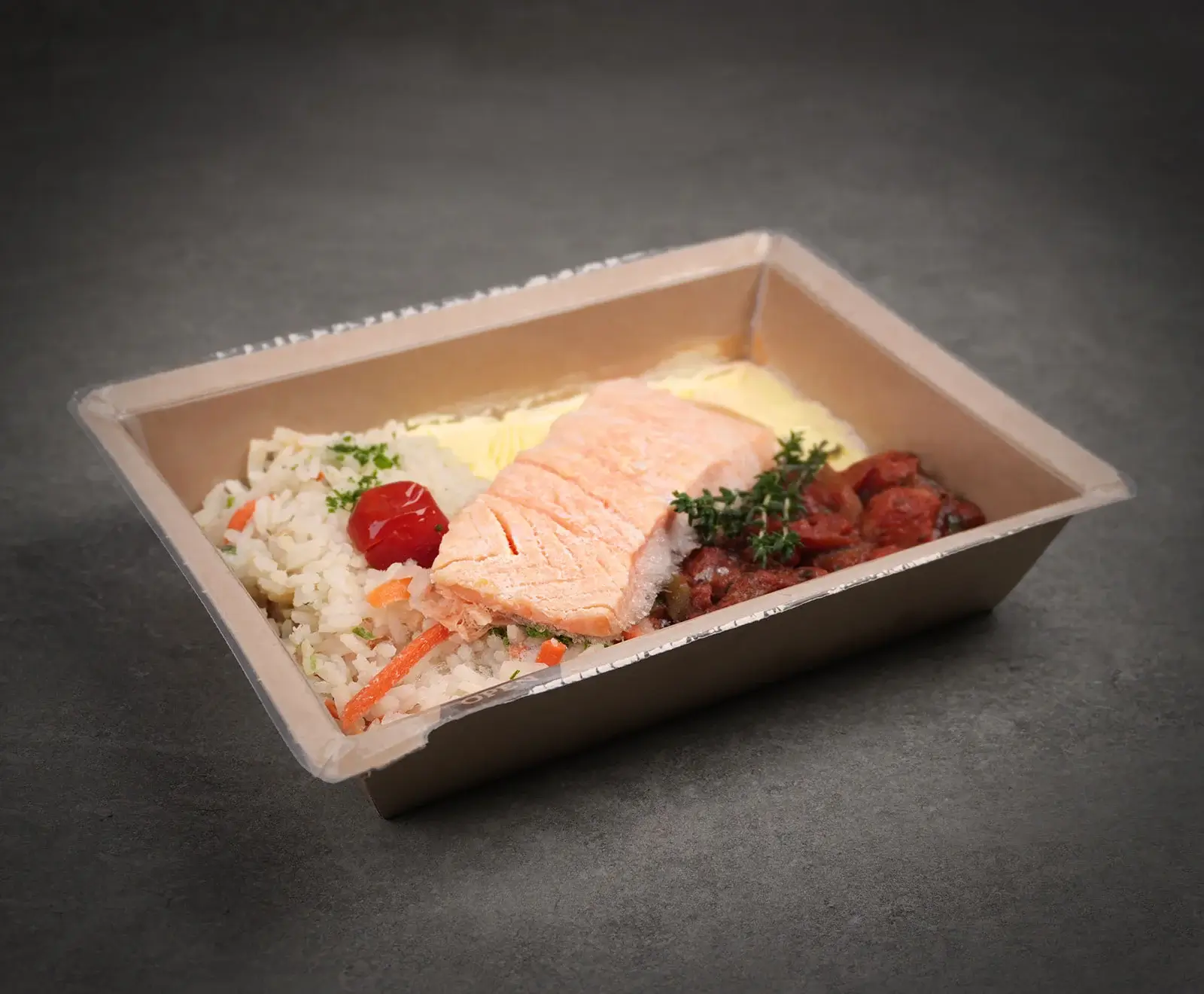 Ein Fertiggericht in einer braunen Pappschale mit transparentem Deckel. Es enthält Lachsfilet, Reis mit Karotten, gelbes Püree und geschmortes Gemüse mit Thymian und Kirschtomate.